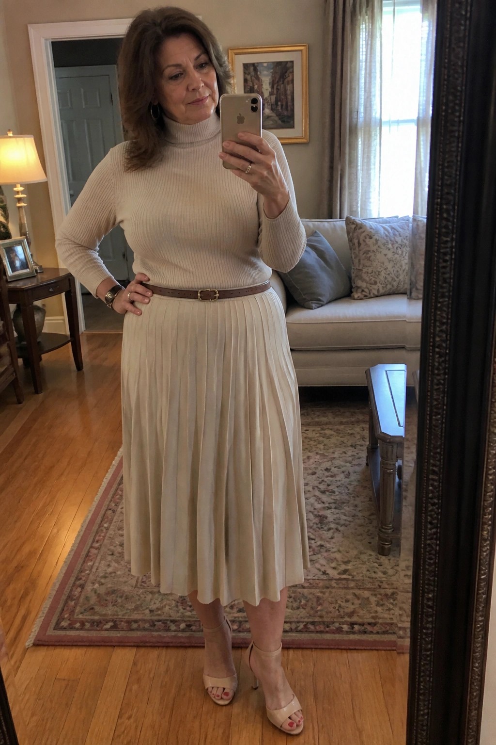 Woman in beige turtleneck and pleated skirt taking mirror selfie in living room