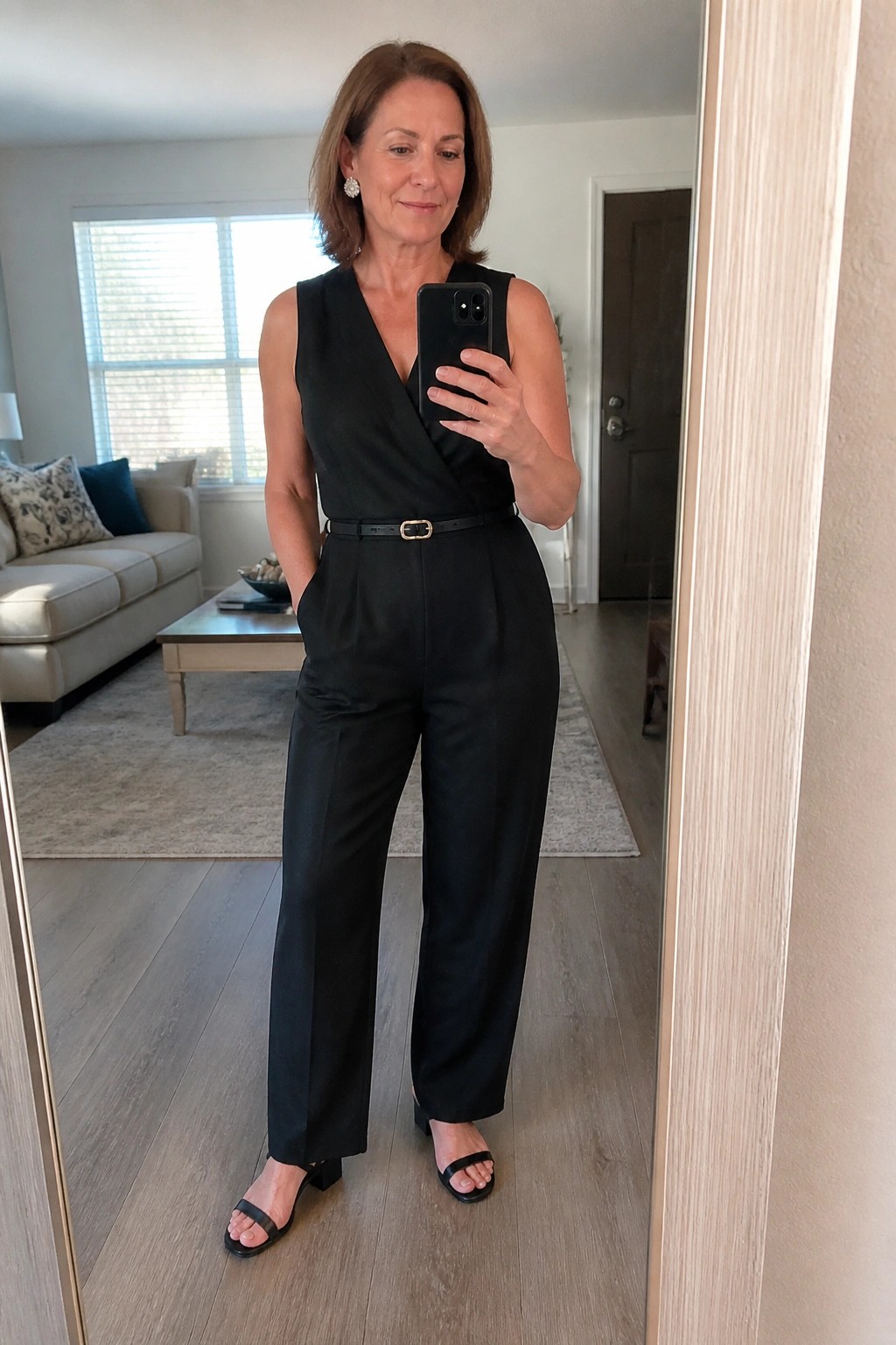 Woman in black belted jumpsuit taking mirror selfie in living room