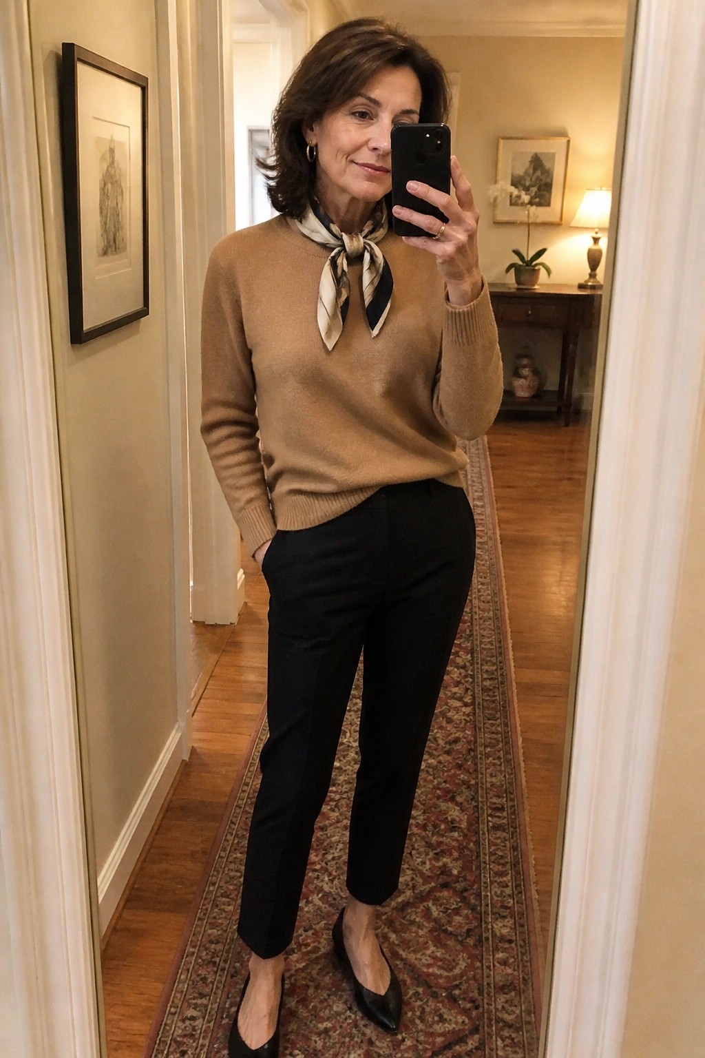 Woman in tan sweater, black pants, and patterned scarf taking mirror selfie in hallway