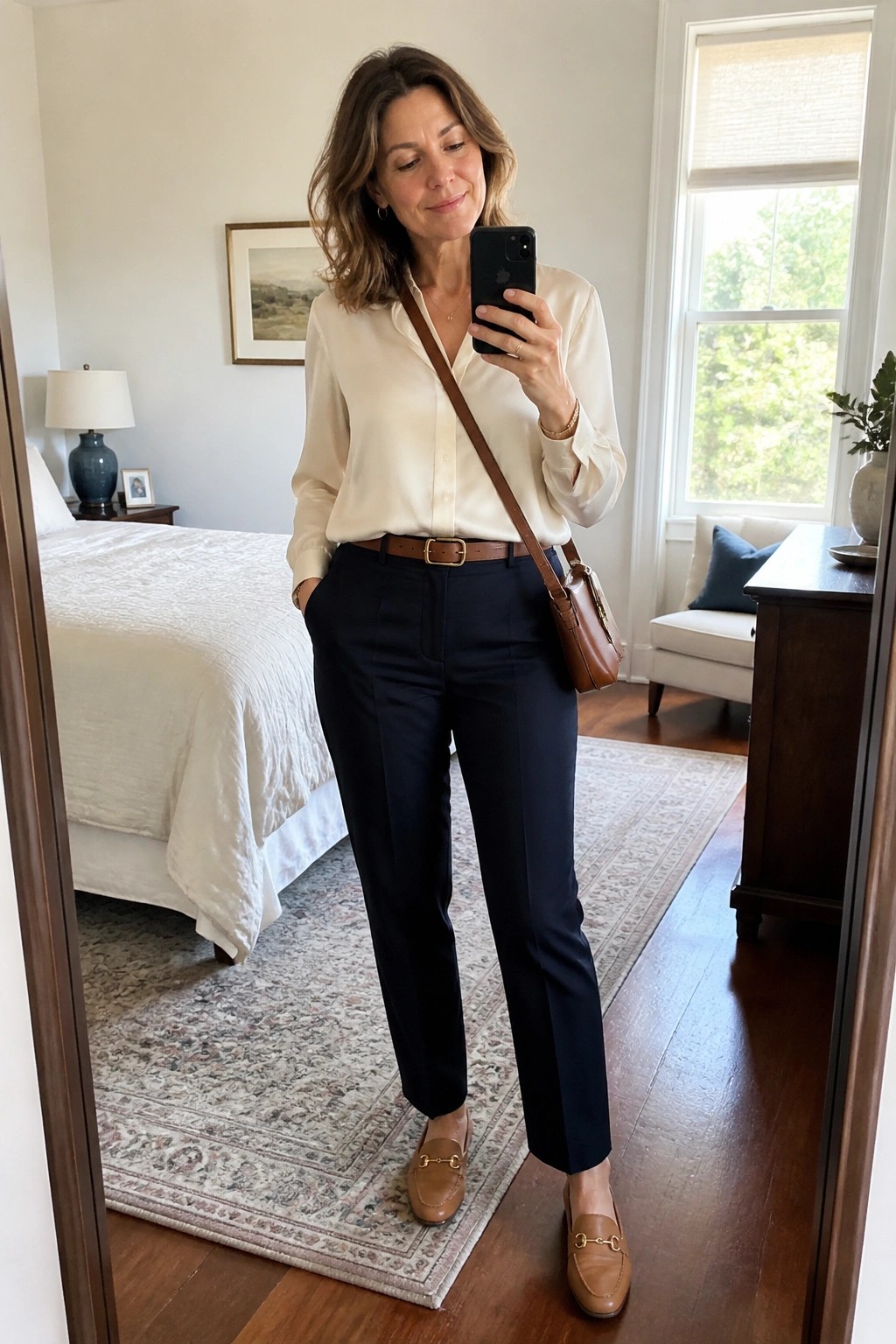 Woman in cream blouse and navy pants taking mirror selfie in bedroom