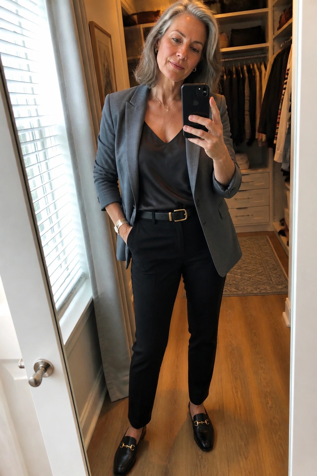 Gray-haired woman in gray blazer, black satin top and pants taking mirror selfie in closet