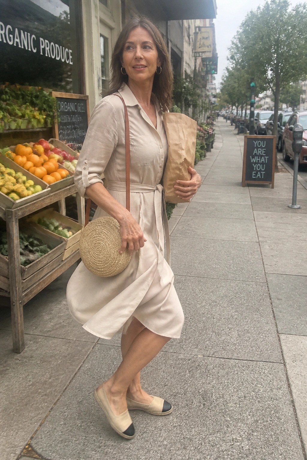 Woman in beige dress holding paper bag and straw purse by organic produce stand