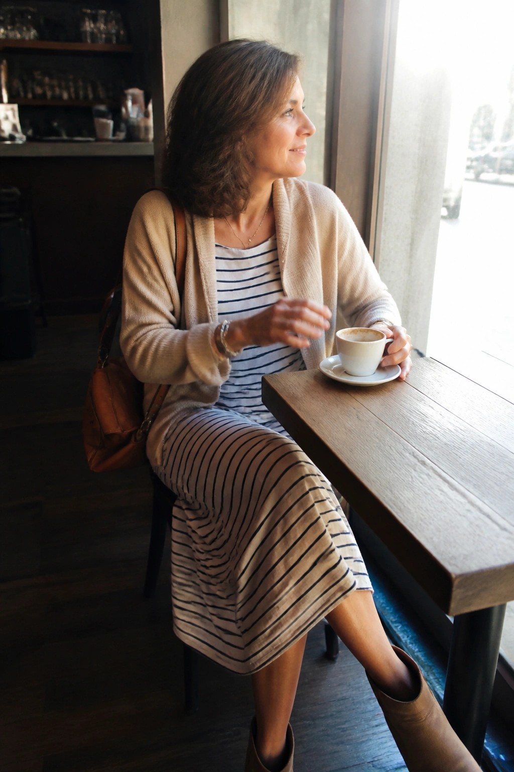 Woman in striped dress and cardigan holding coffee at cafe window table