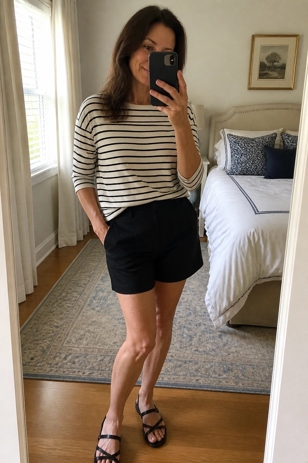 Woman in her fifties wearing a black and white striped three quarter sleeve top tucked loosely into black shorts with strappy black flat sandals while taking a mirror selfie