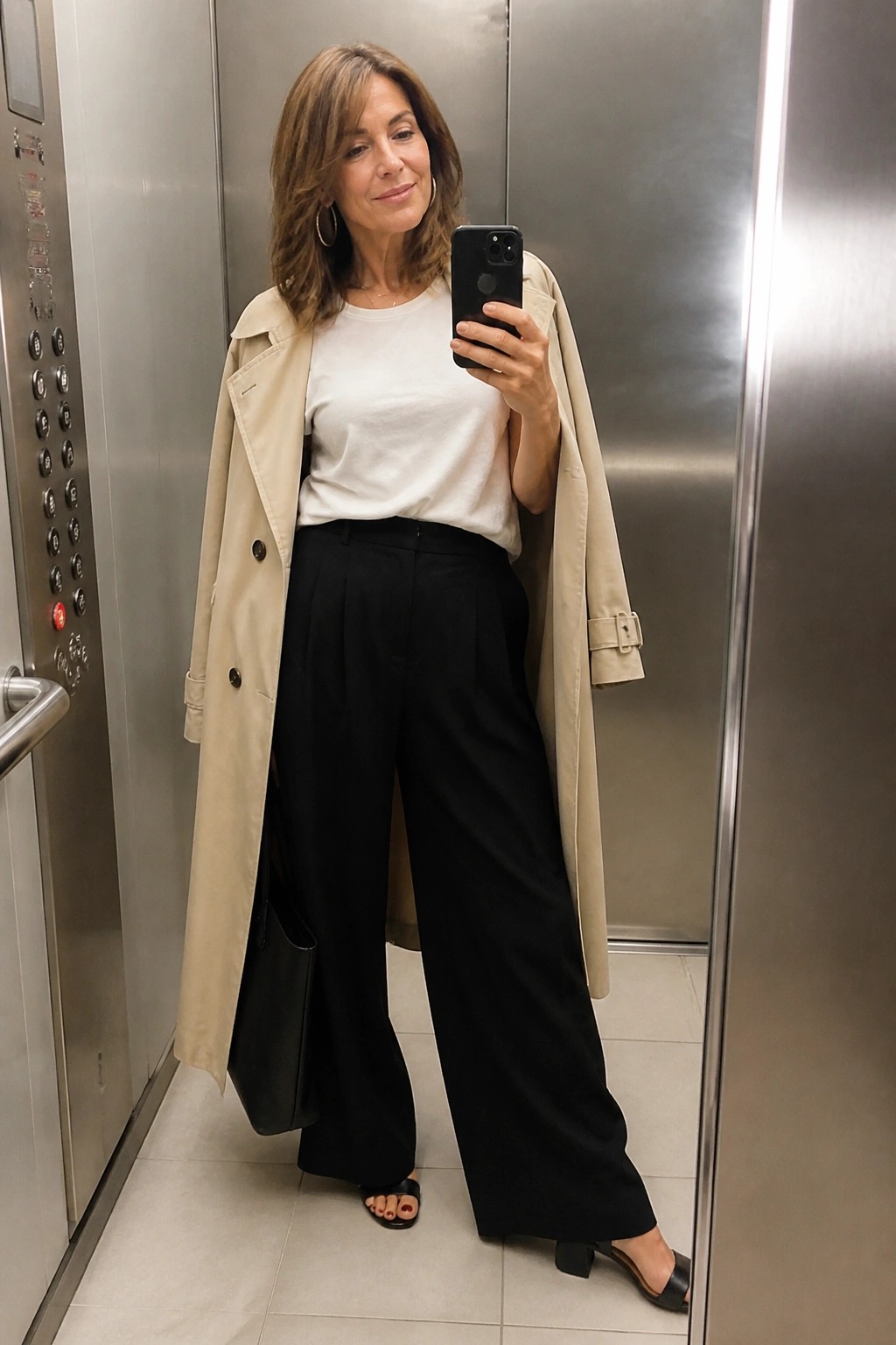 Woman in beige trench coat and black wide-leg pants taking selfie in elevator