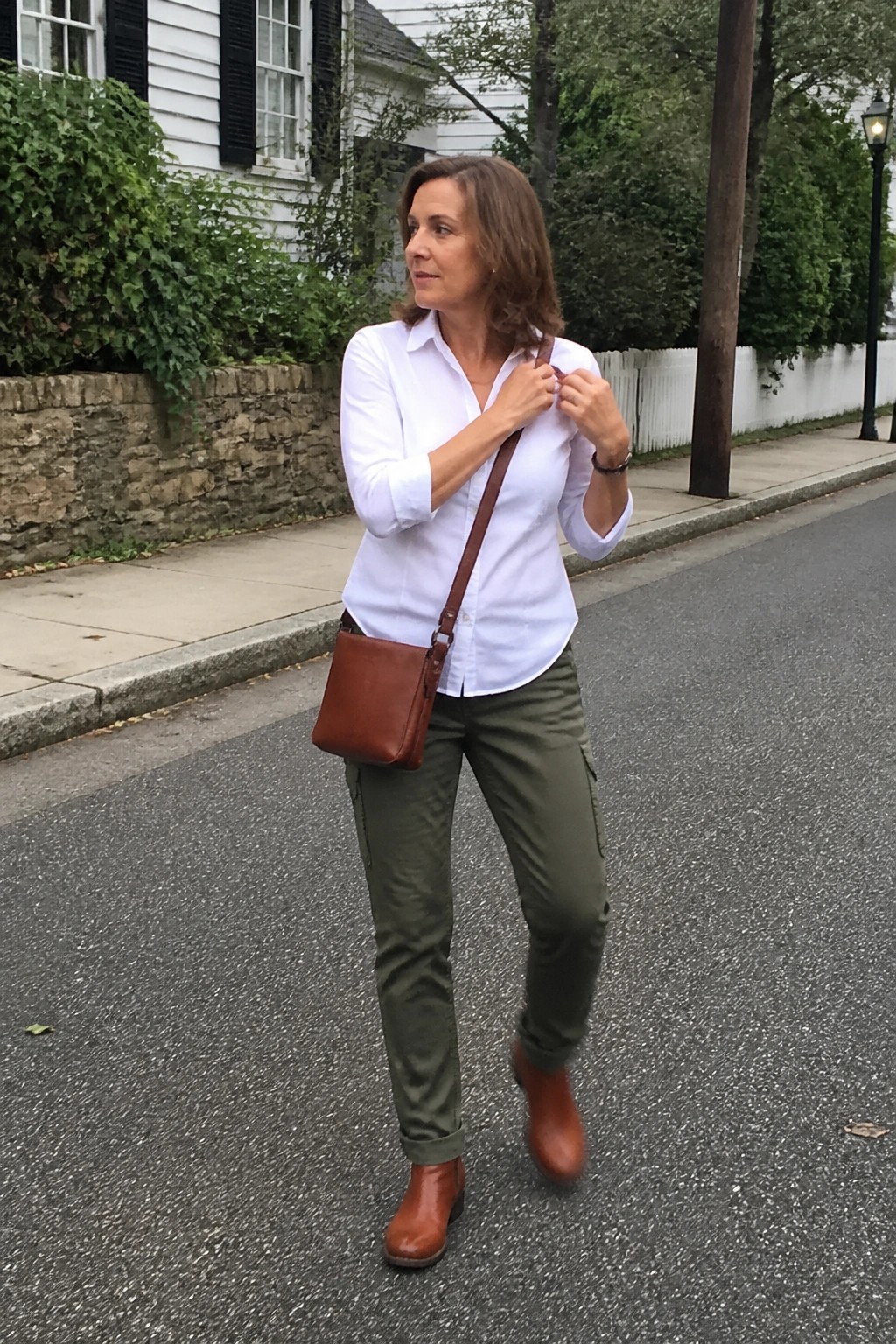 Woman in white shirt and green pants adjusts brown leather crossbody bag on street
