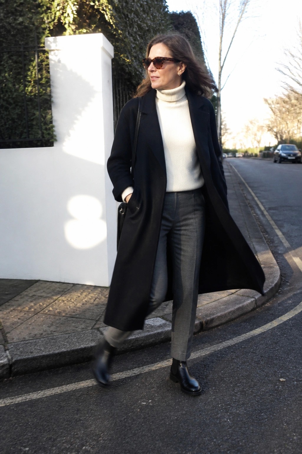 Woman in black coat and white turtleneck walks on sunny urban street.
