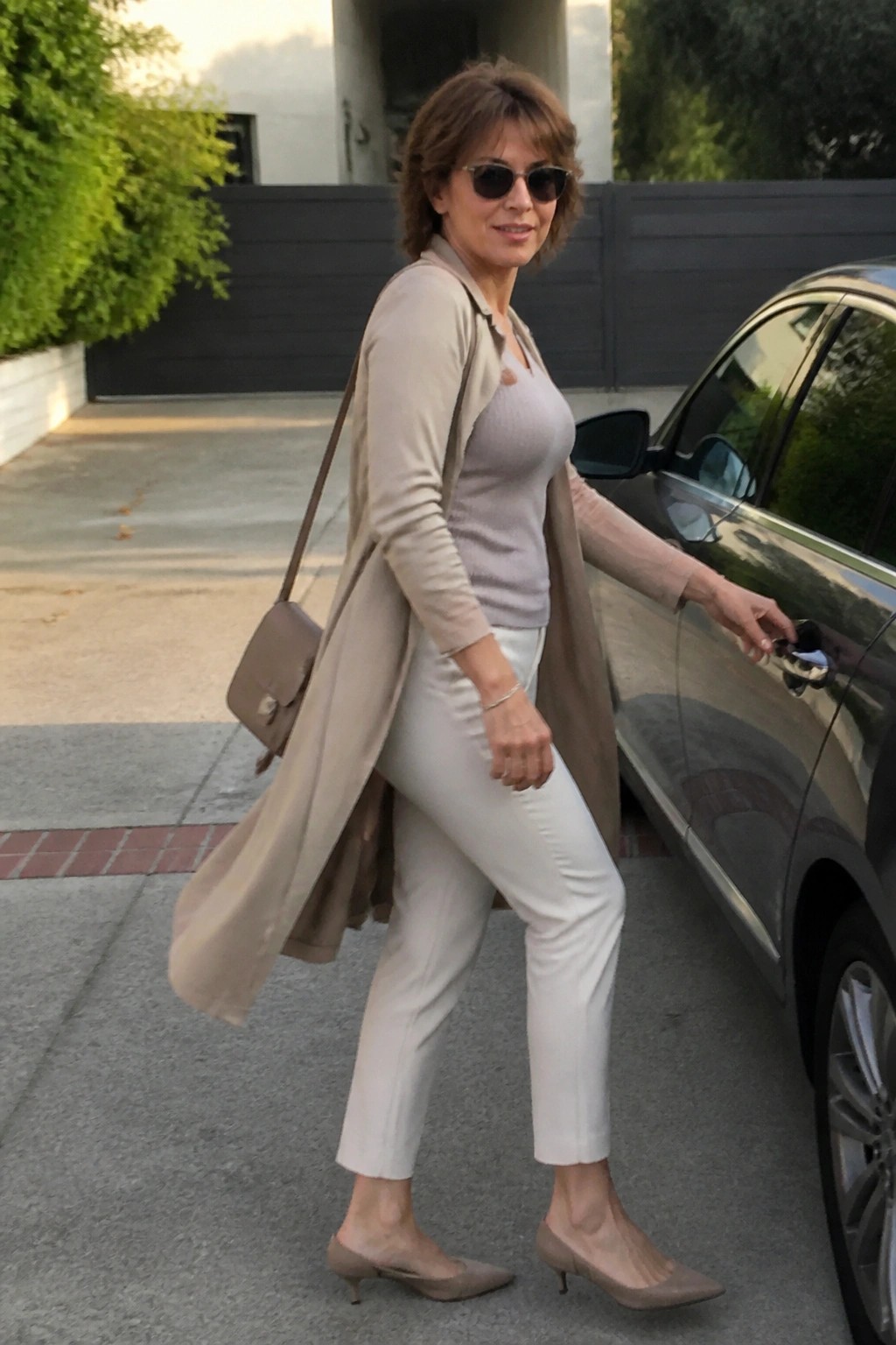 Woman in beige cardigan and white pants opening dark car door outdoors