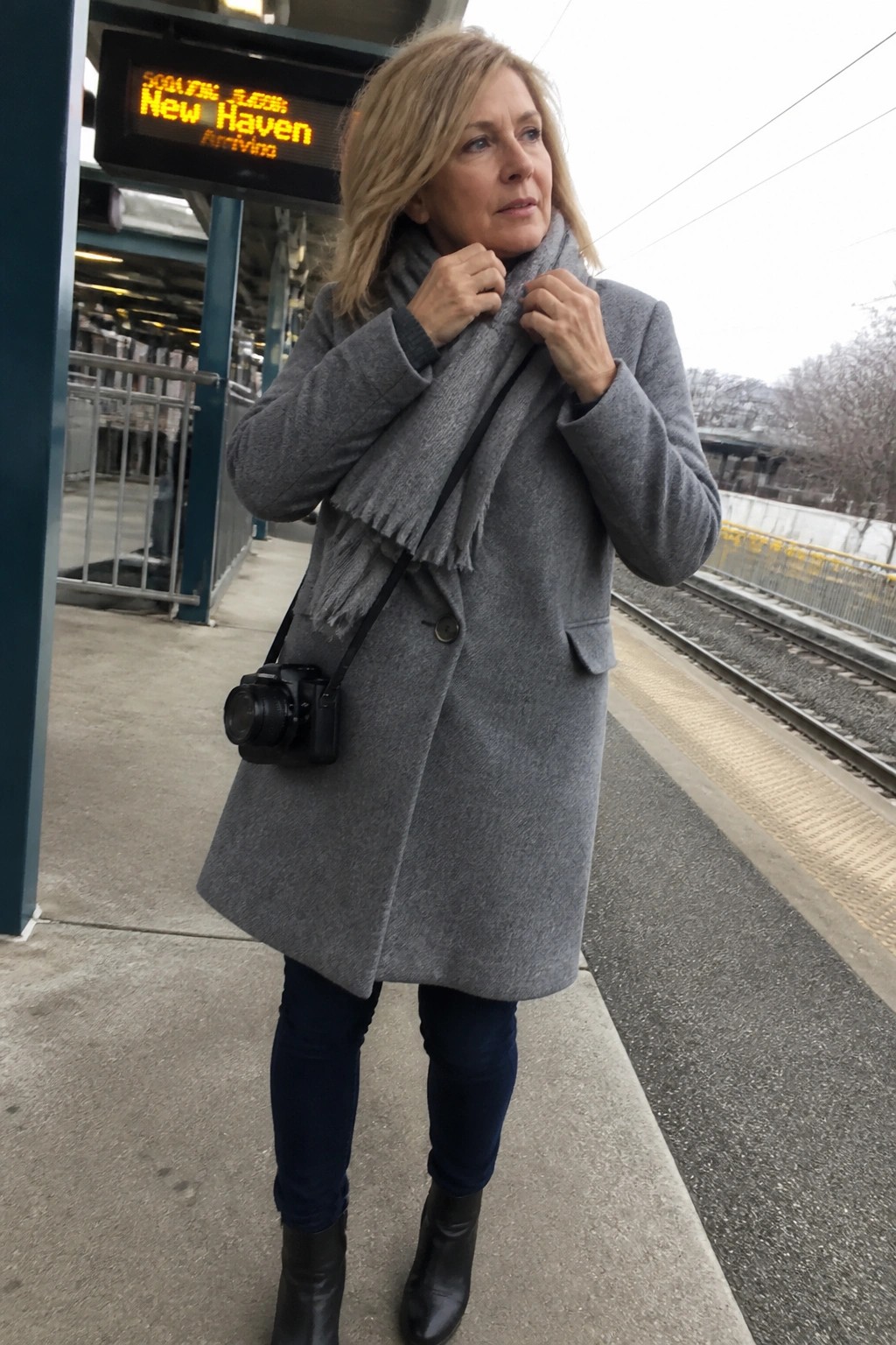 Blonde woman in gray coat adjusts scarf on train platform with camera
