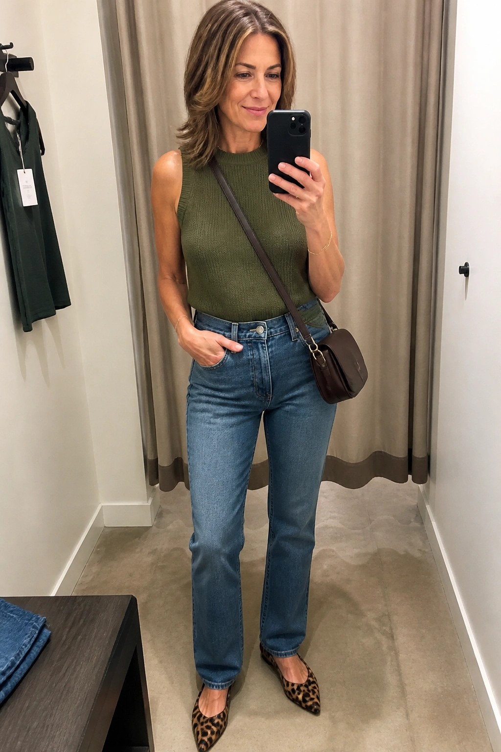 Woman in olive green sleeveless top and blue jeans taking mirror selfie in fitting room