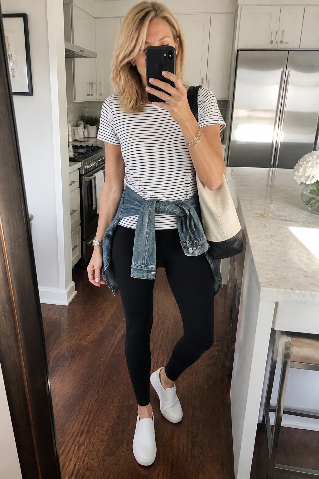 Woman wearing black and white striped short sleeve top with black leggings denim jacket tied at waist white slip on sneakers and carrying a beige tote bag while taking a mirror selfie in a kitchen.