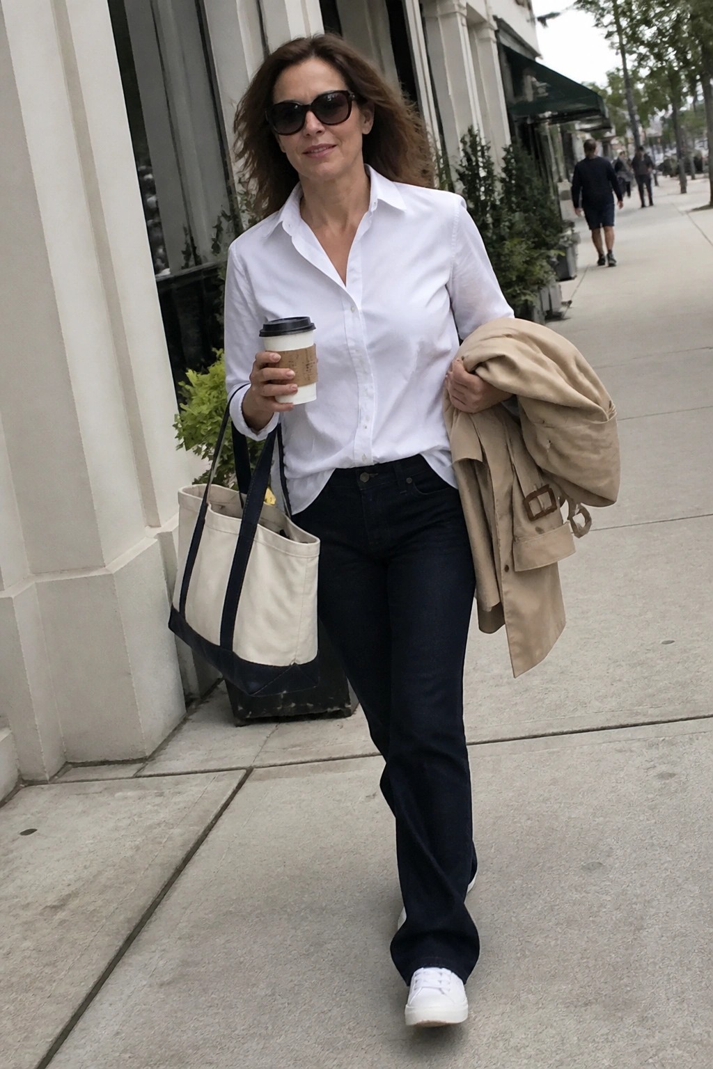 Woman in white shirt and jeans carrying coffee, tote bag, and trench coat on sidewalk