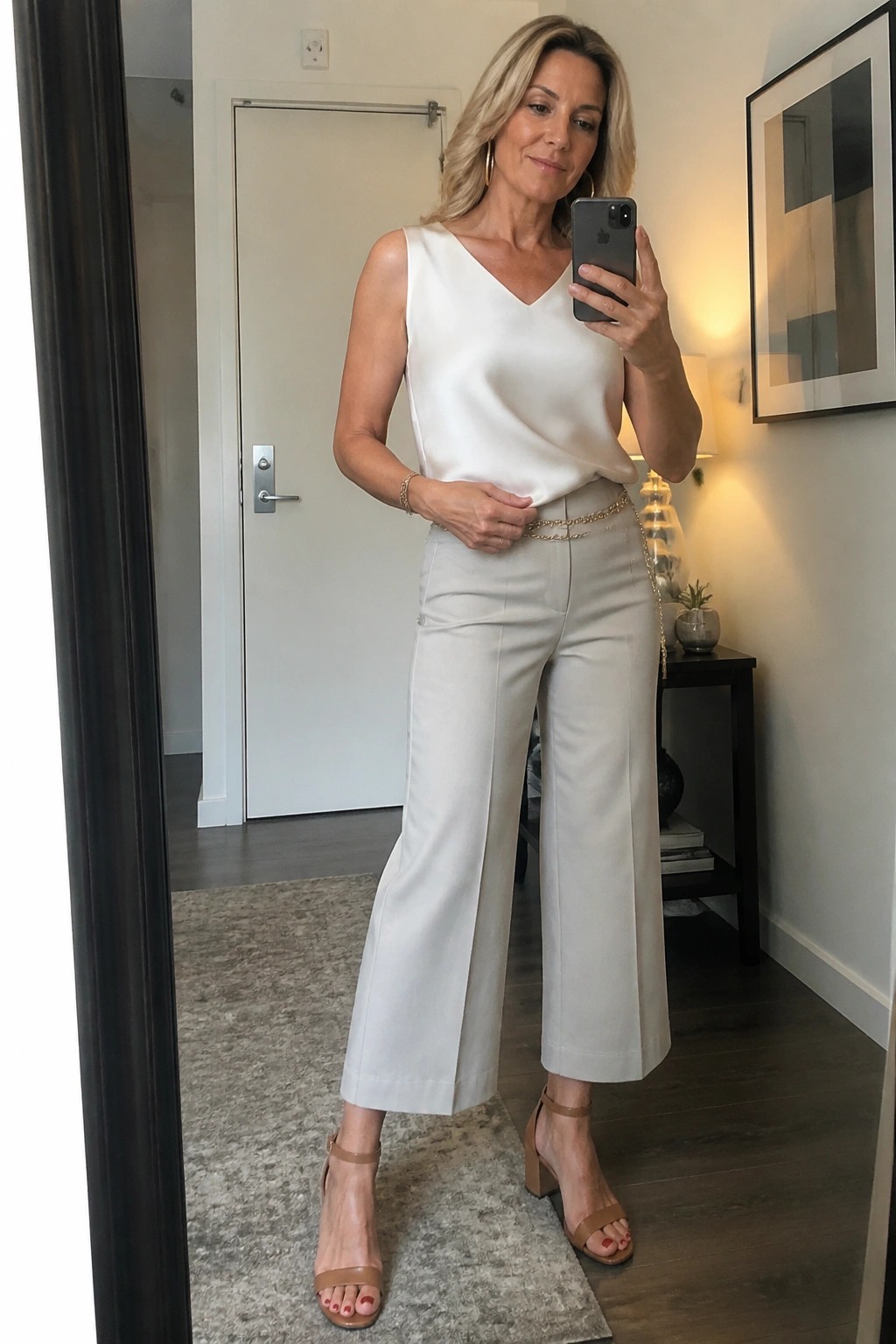 Woman over 50 in a white sleeveless V-neck top tucked into light beige wide-leg cropped pants with a thin gold chain belt, paired with nude block heel sandals and gold hoop earrings, taking a mirror selfie.
