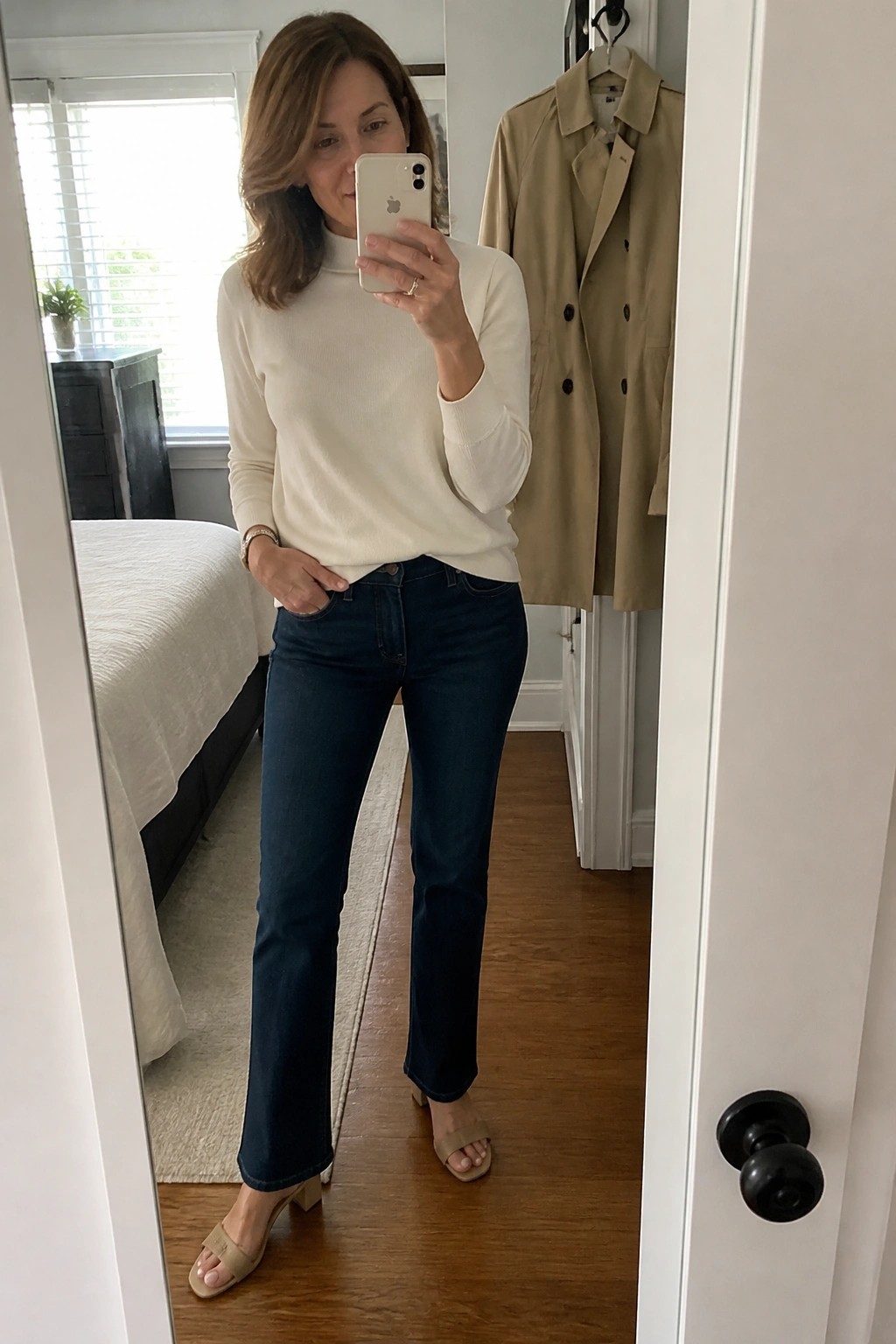 Woman in white turtleneck sweater and dark jeans taking bedroom mirror selfie