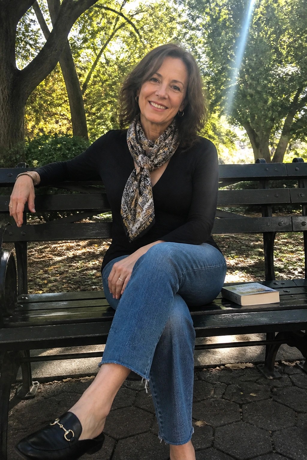 Smiling woman in black top, scarf, and jeans on sunny park bench beside book