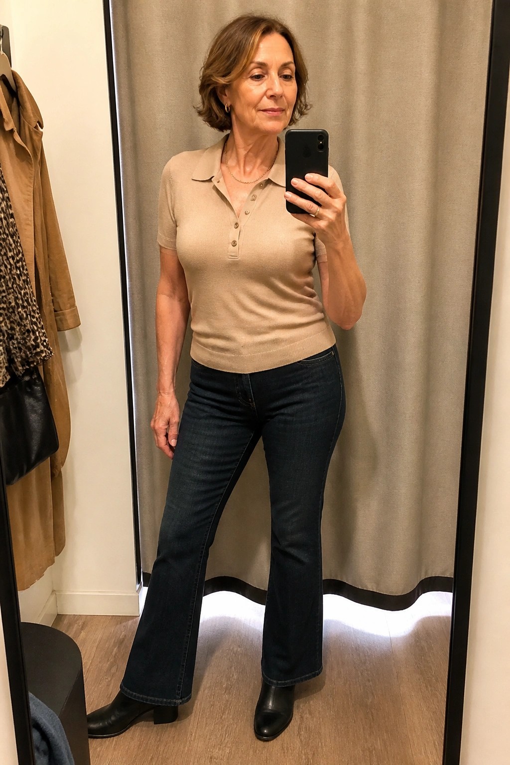 Woman in beige polo sweater and dark flared jeans taking mirror selfie in fitting room