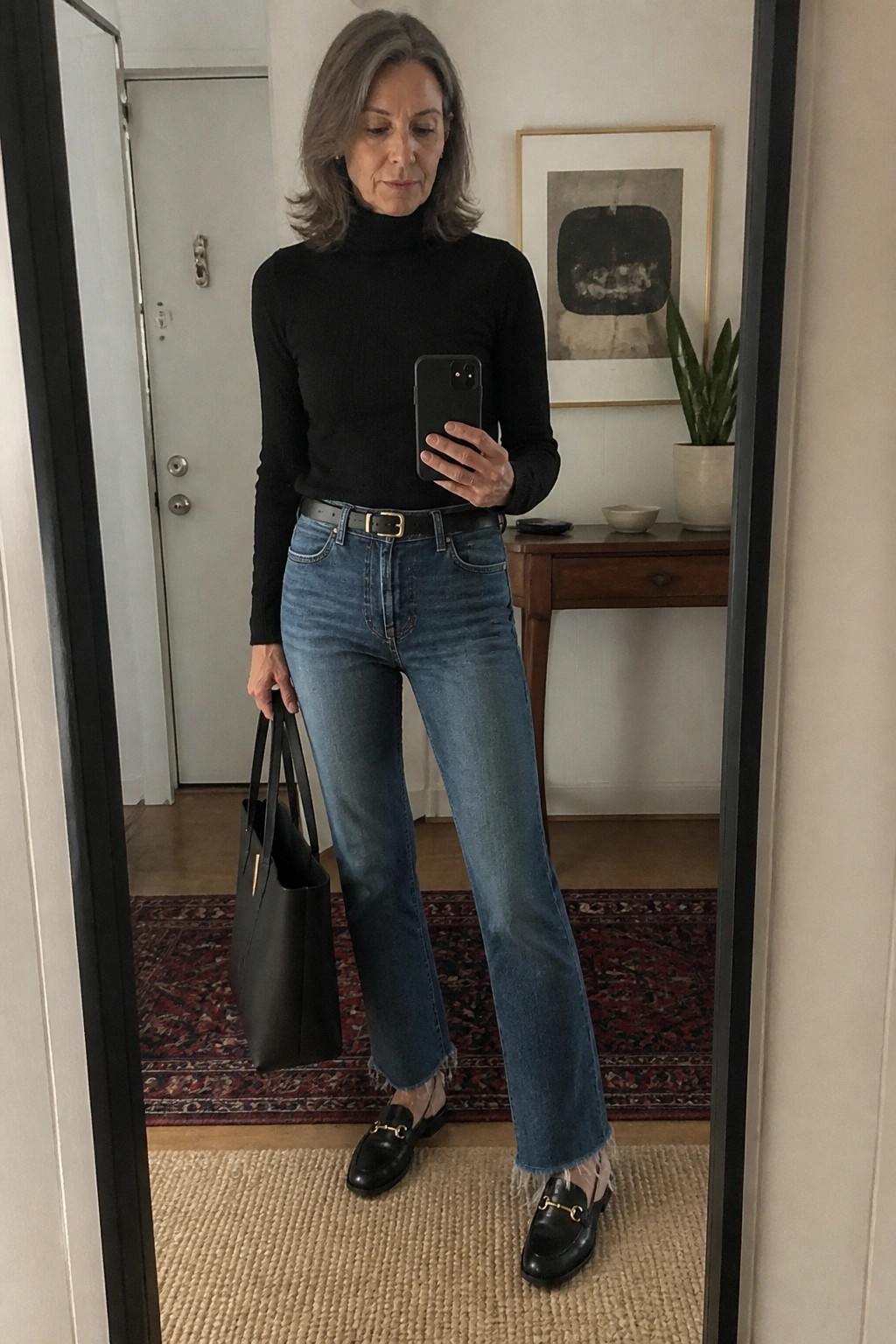 Woman in black turtleneck and blue jeans holds phone and tote in mirror selfie