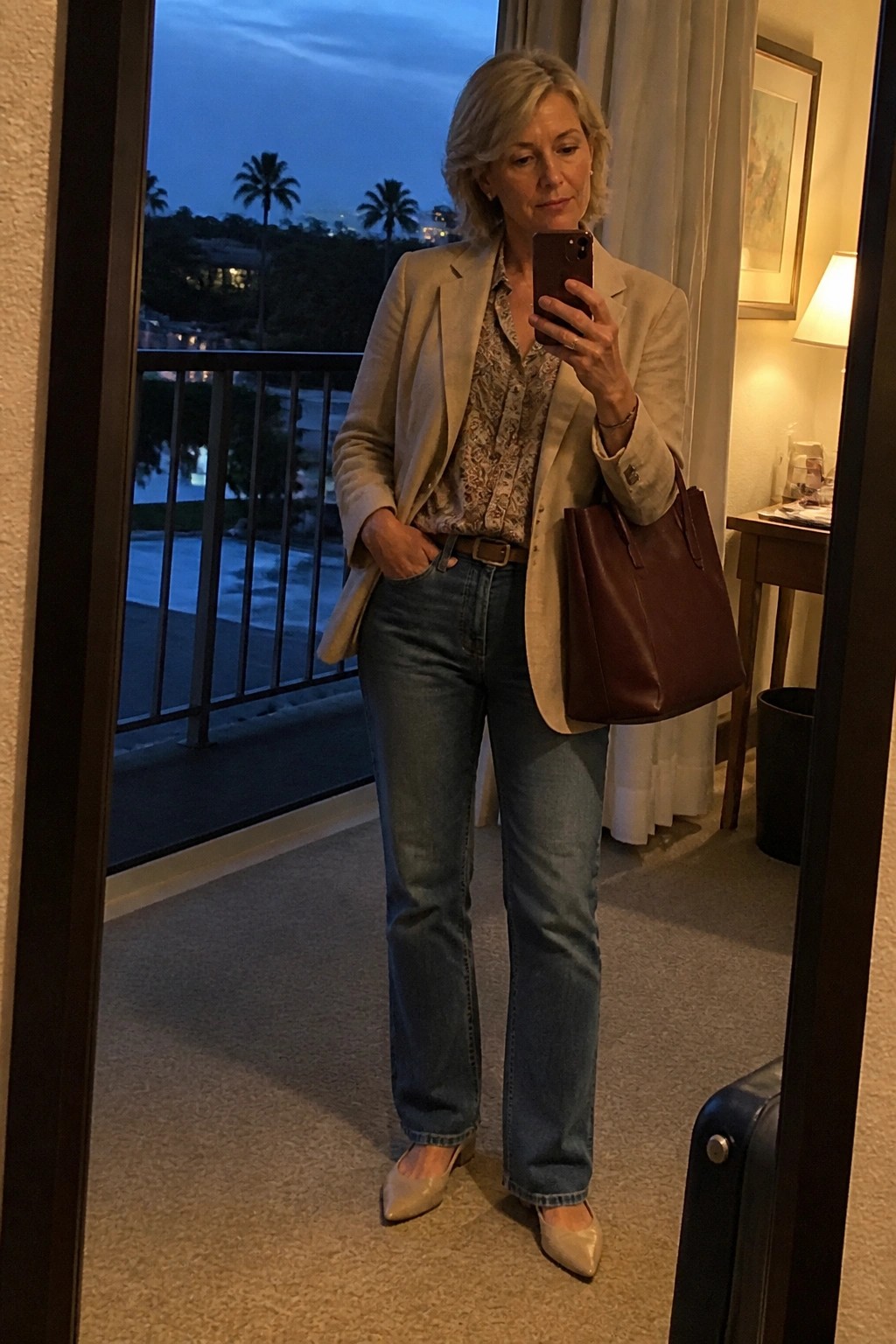 Woman in beige blazer and jeans taking mirror selfie with brown leather tote at dusk