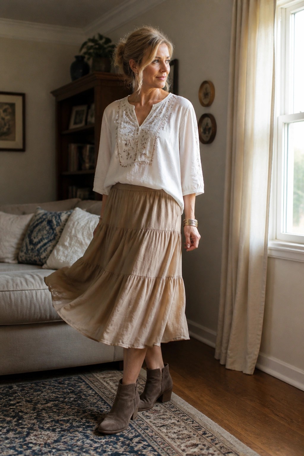 A woman over 50 in a white embroidered blouse over a beige tiered midi skirt paired with gray suede ankle boots and simple bracelets.