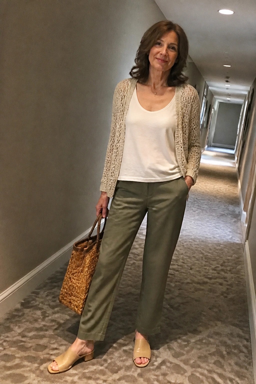 Woman in beige knit cardigan, white tank, green pants holding woven tote in hallway