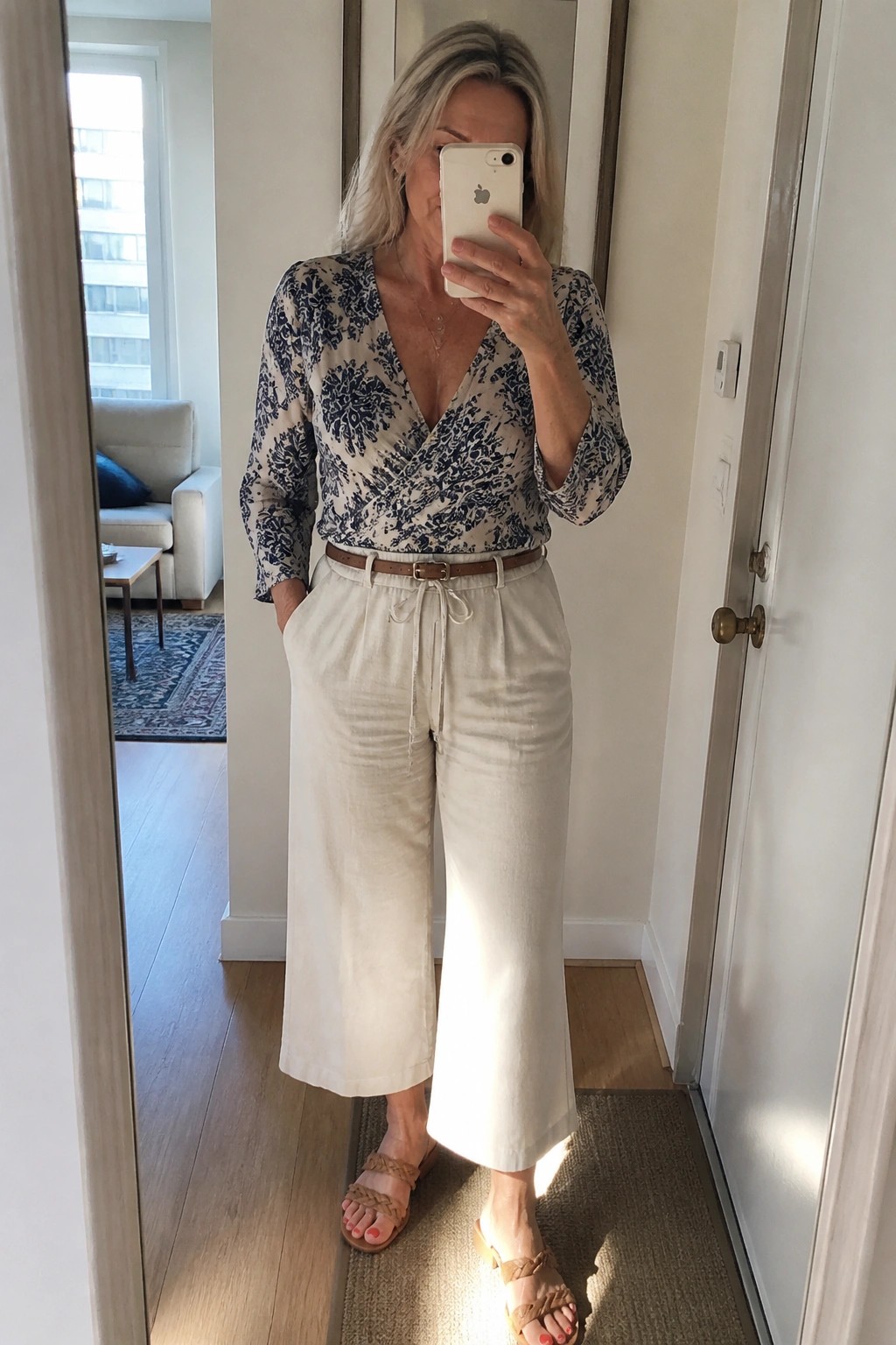 Woman in her fifties wearing a blue and white floral wrap top tucked into cream wide leg linen pants with a brown belt and braided sandals, taking a mirror selfie.