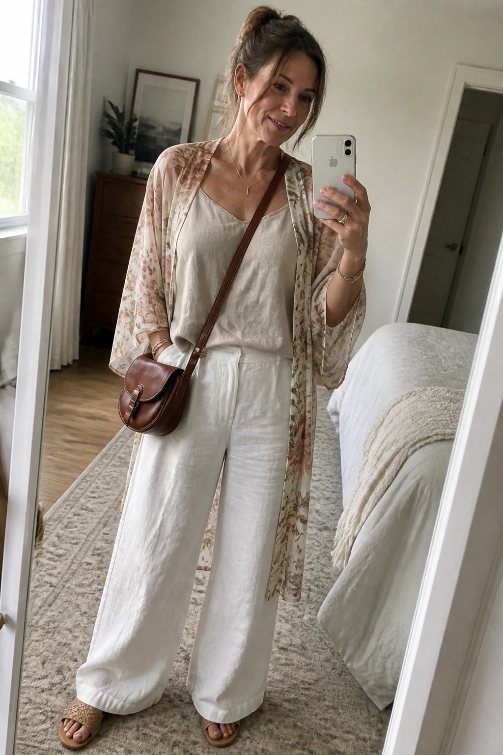 Woman in white linen and floral kimono taking mirror selfie with brown bag.
