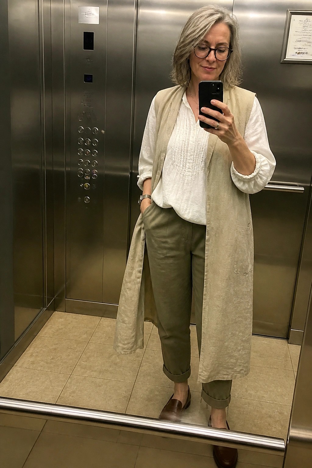 Gray-haired woman in beige vest and olive pants taking elevator mirror selfie.