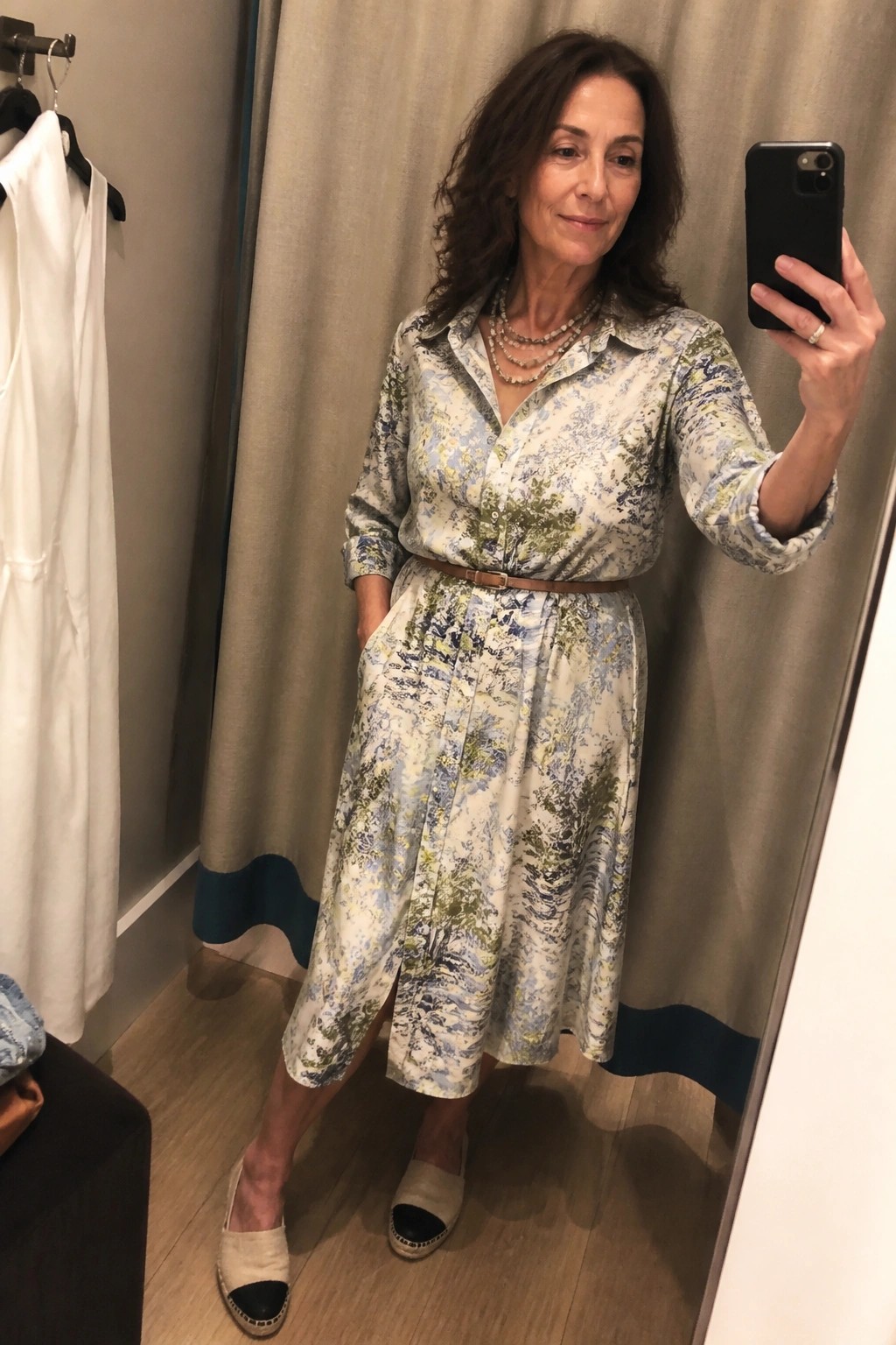 Woman in blue-green patterned shirt dress takes mirror selfie in fitting room