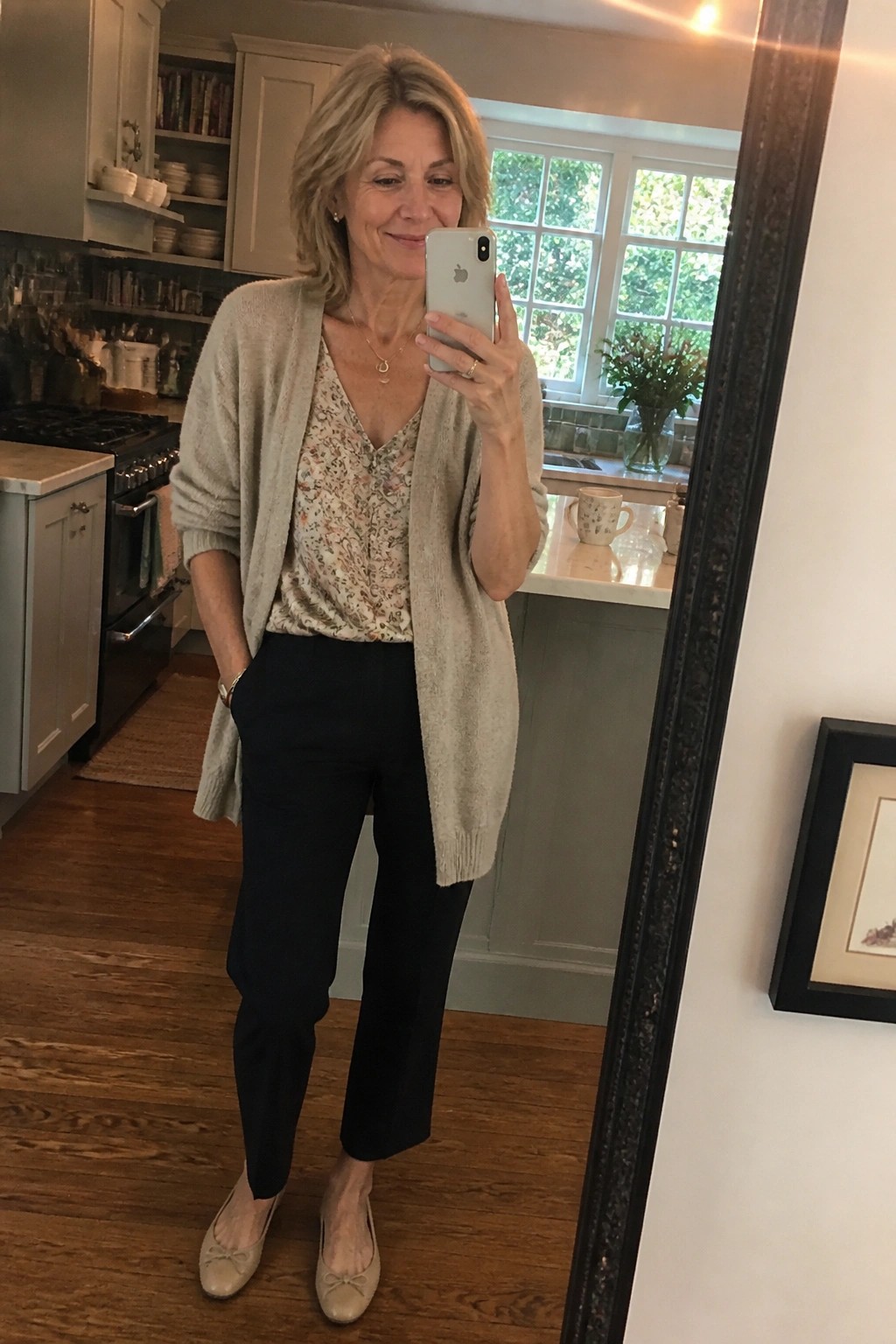 Blonde woman in floral top and beige cardigan taking mirror selfie in kitchen