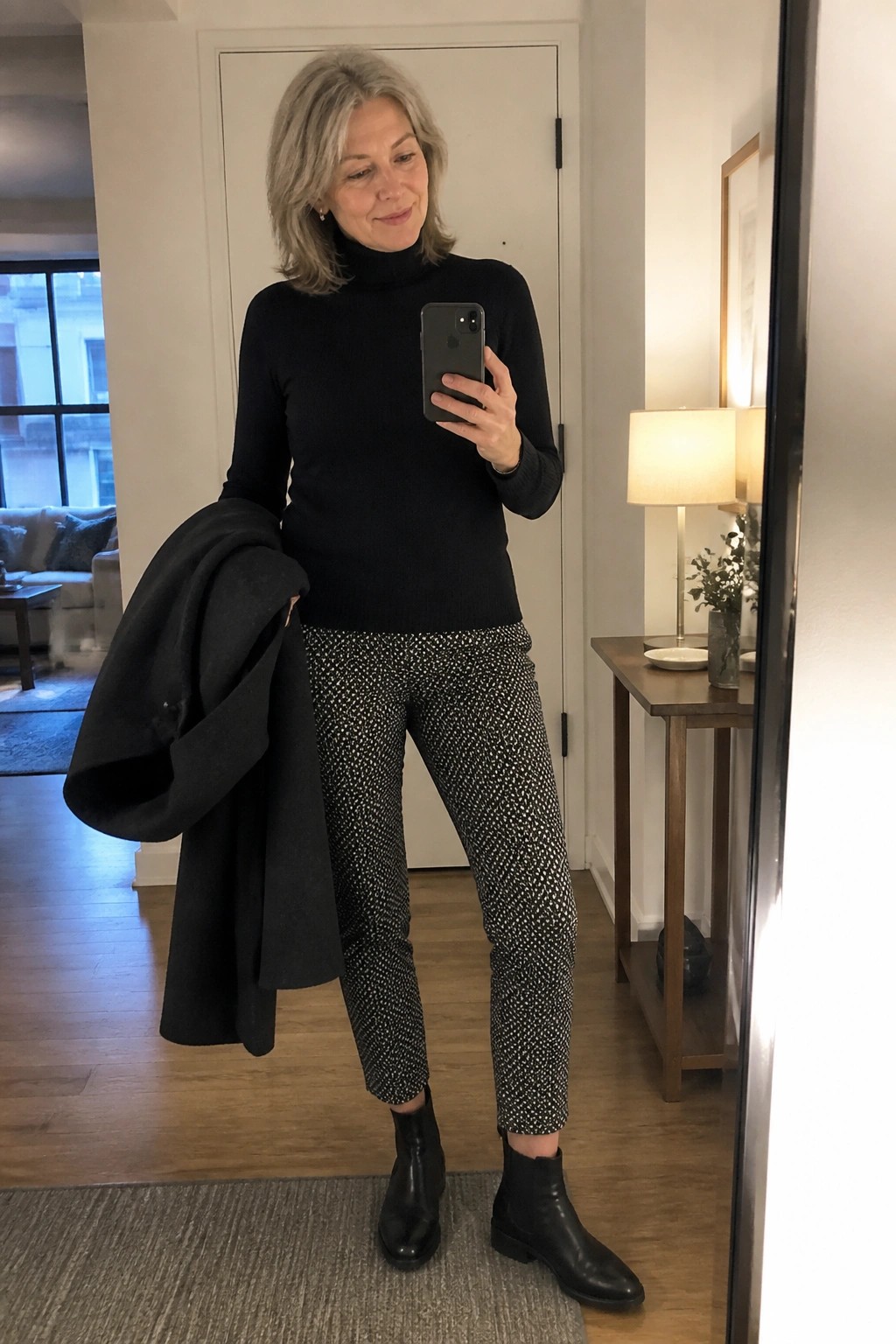 Woman in black turtleneck and patterned pants holds coat while taking mirror selfie.