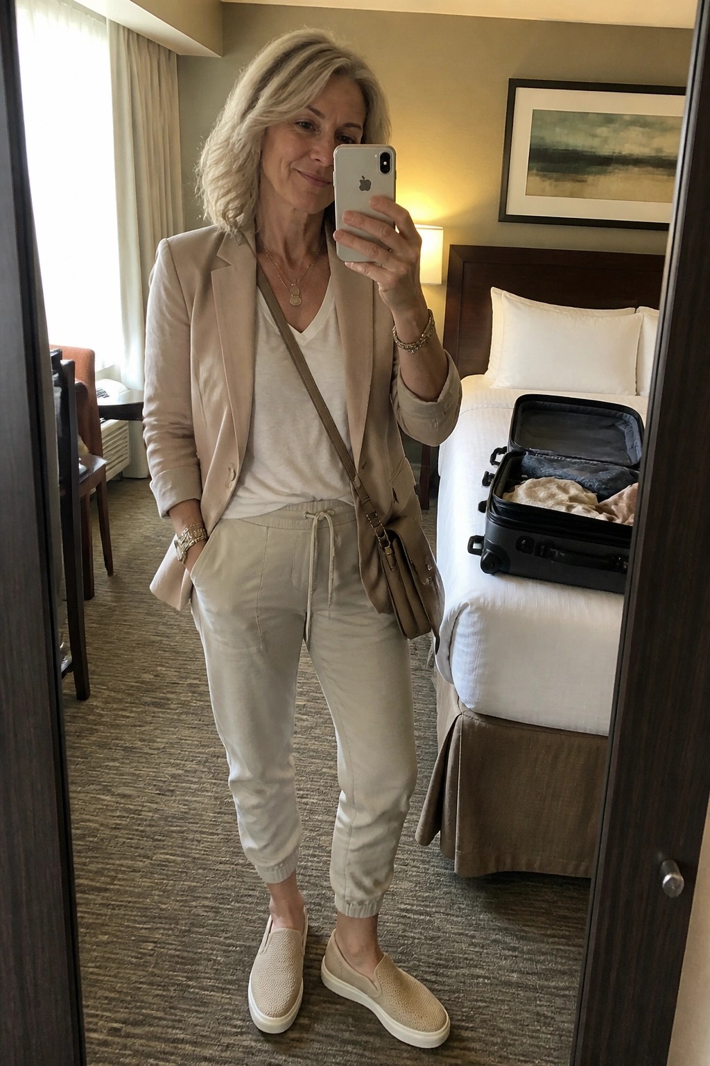 Woman over 50 wearing a beige blazer over a white tee with matching jogger pants, beige slip on sneakers, and a crossbody bag in a hotel room mirror selfie.