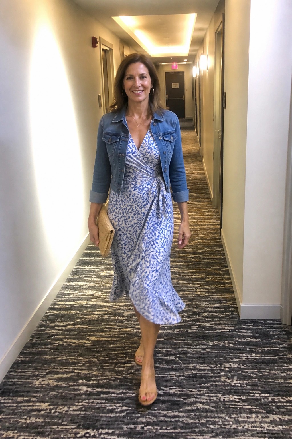 Smiling woman in denim jacket and blue patterned dress walks hotel hallway