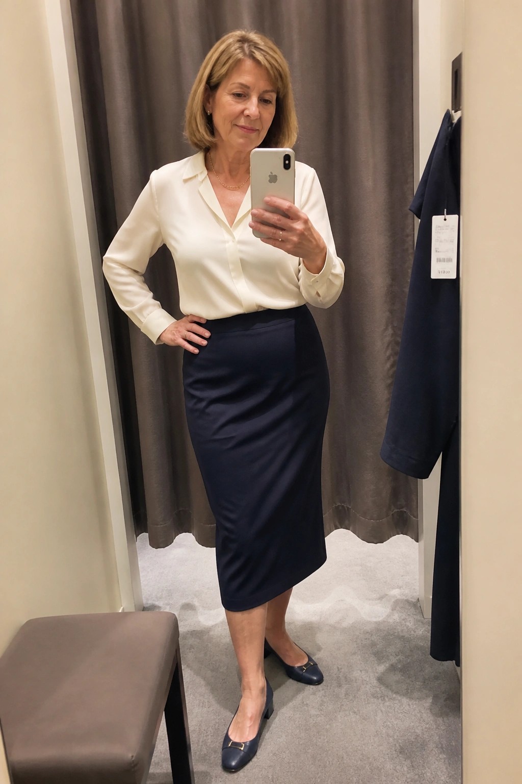 Woman in cream blouse and navy skirt taking mirror selfie in fitting room
