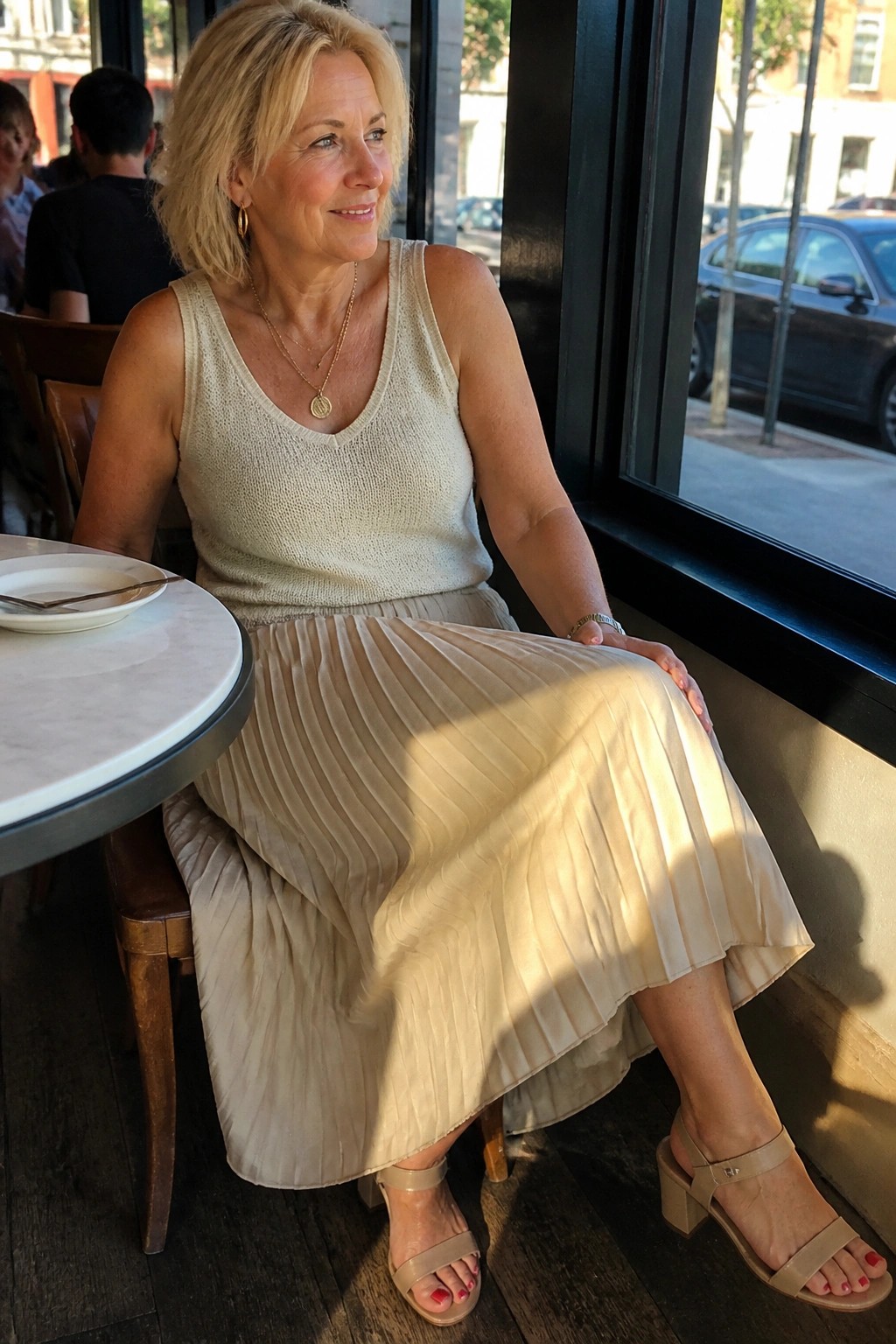 Blonde woman in beige knit top and pleated skirt by sunny cafe window
