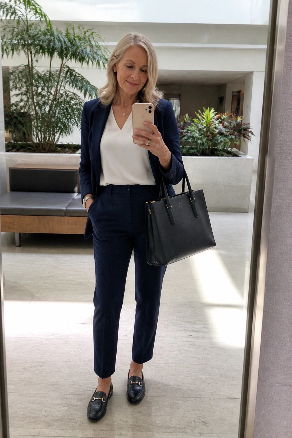 Blonde woman in navy pantsuit taking mirror selfie with black handbag in modern lobby