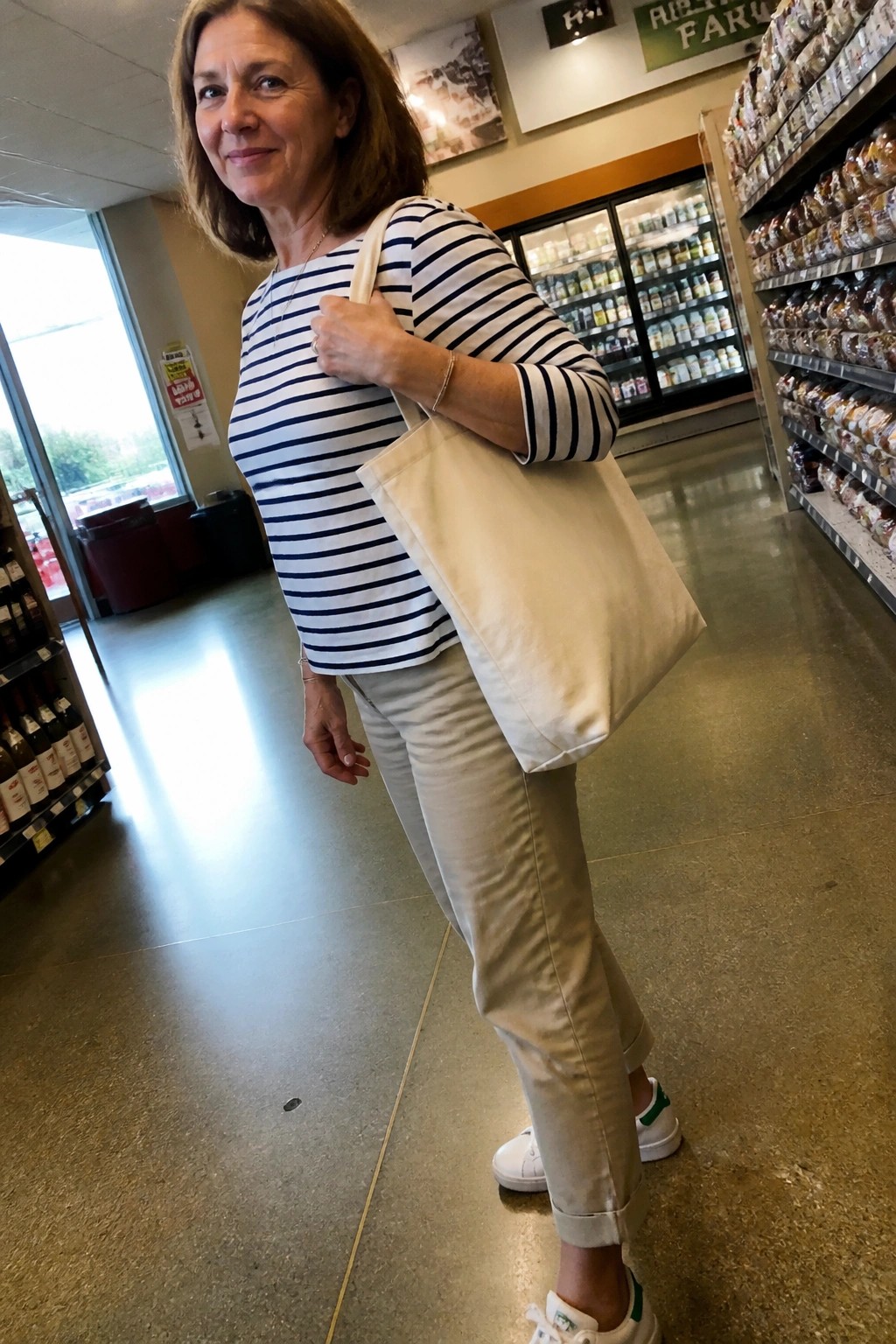 A woman over 50 in a navy and white striped shirt tucked into beige pants with white sneakers and a beige tote bag standing in a store aisle.
