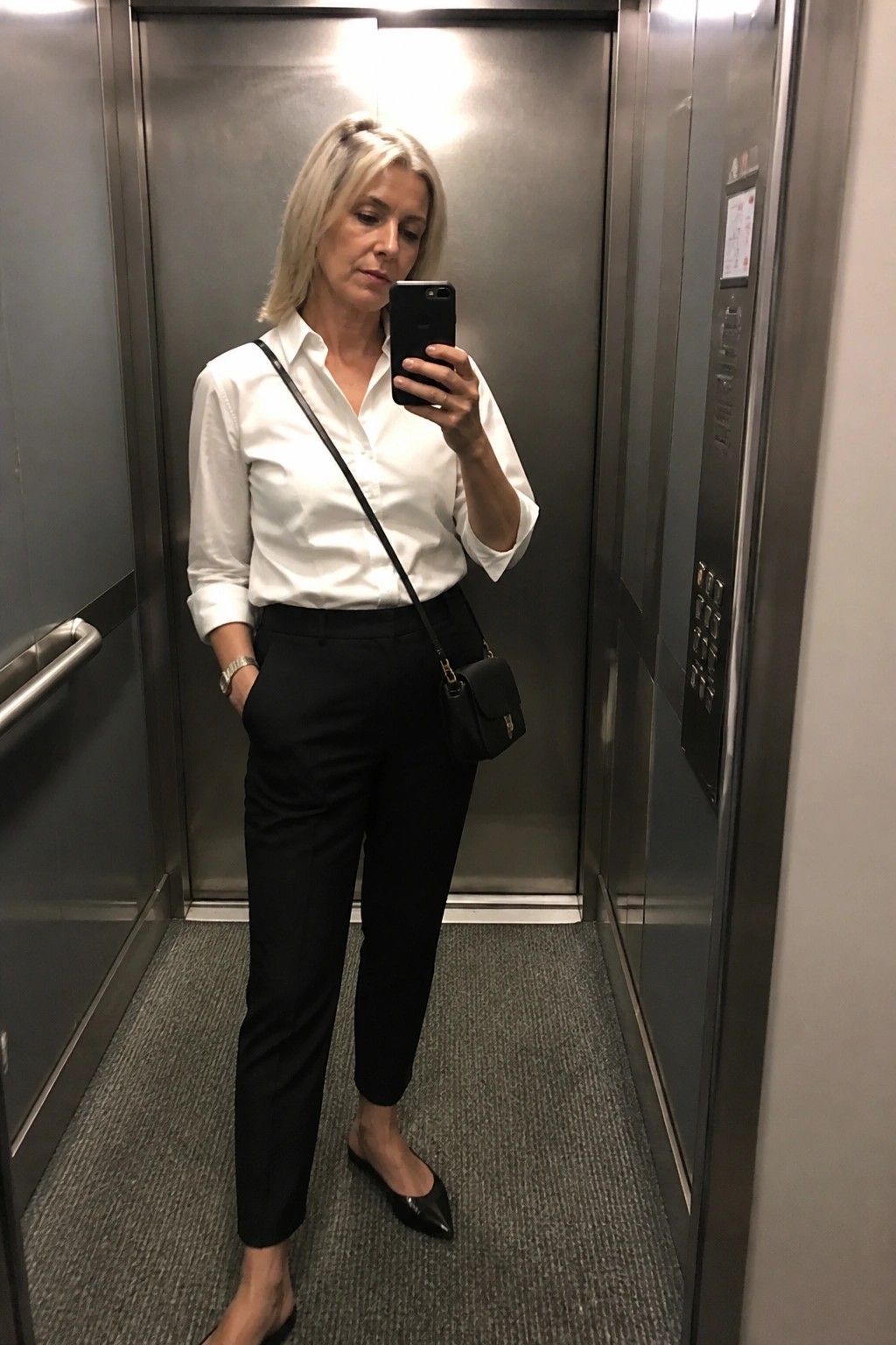 Blonde woman in white shirt and black pants taking selfie in elevator