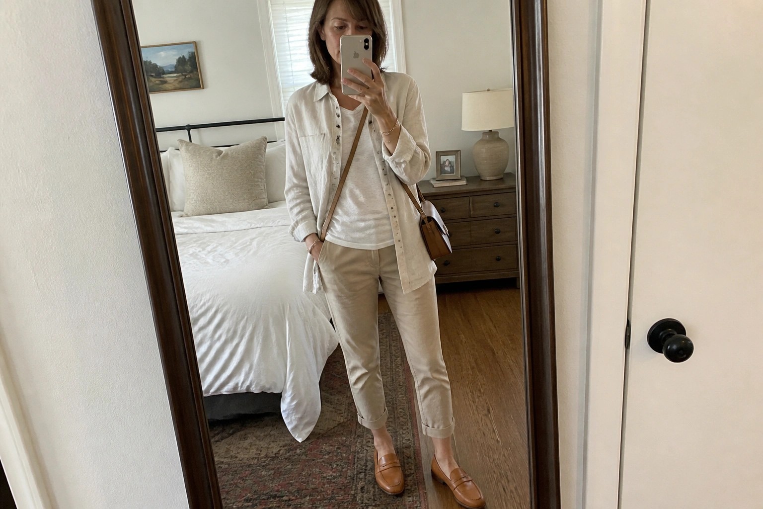 Woman in white shirt and beige pants taking mirror selfie in bedroom