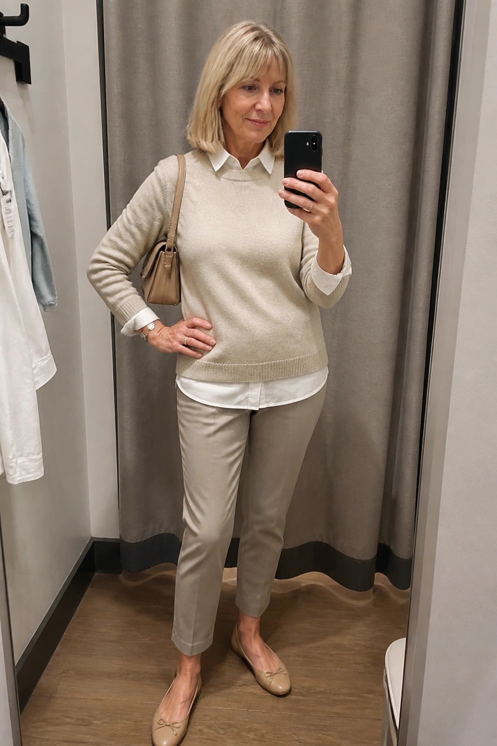 Woman in beige sweater over white shirt taking mirror selfie in fitting room