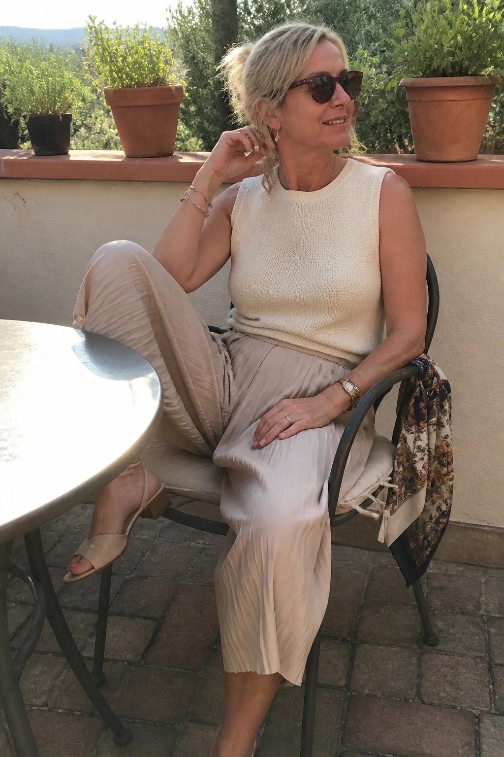 Blonde woman in cream knit top and pleated beige pants on sunny patio chair