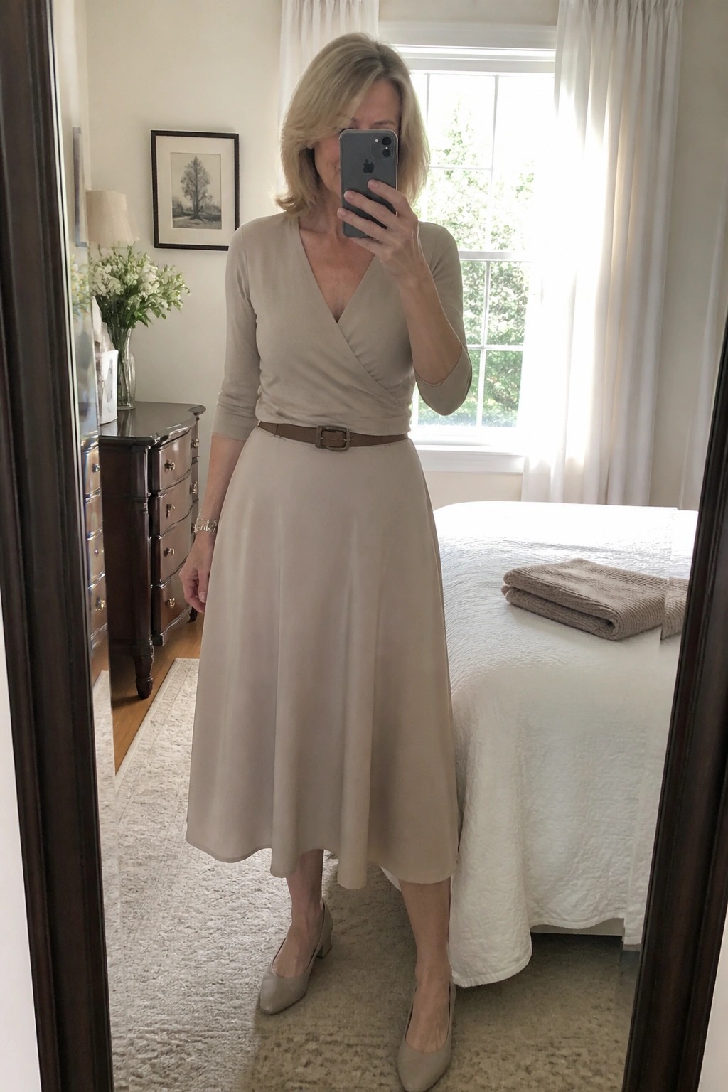 Woman in beige wrap top and midi skirt taking mirror selfie in bedroom