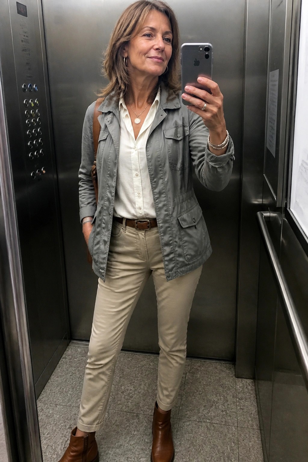 Woman in gray utility jacket and beige pants taking selfie in elevator.