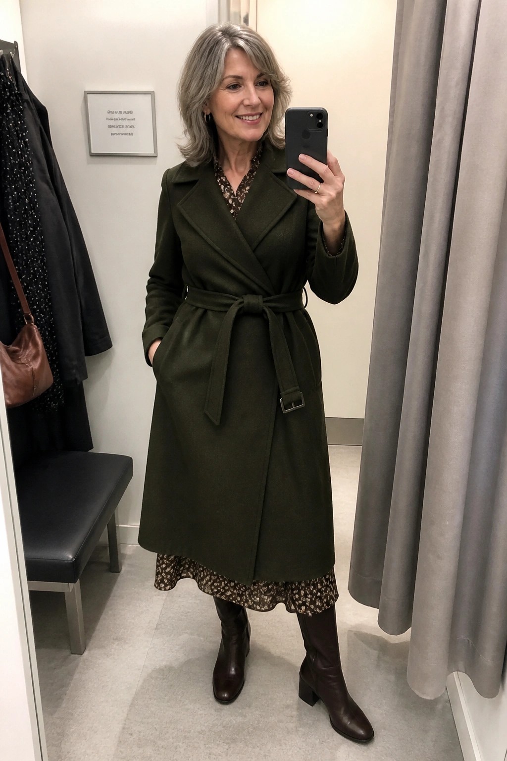 Woman over 50 in olive green belted coat layered over floral print dress paired with brown knee high boots taking a mirror selfie in a fitting room.