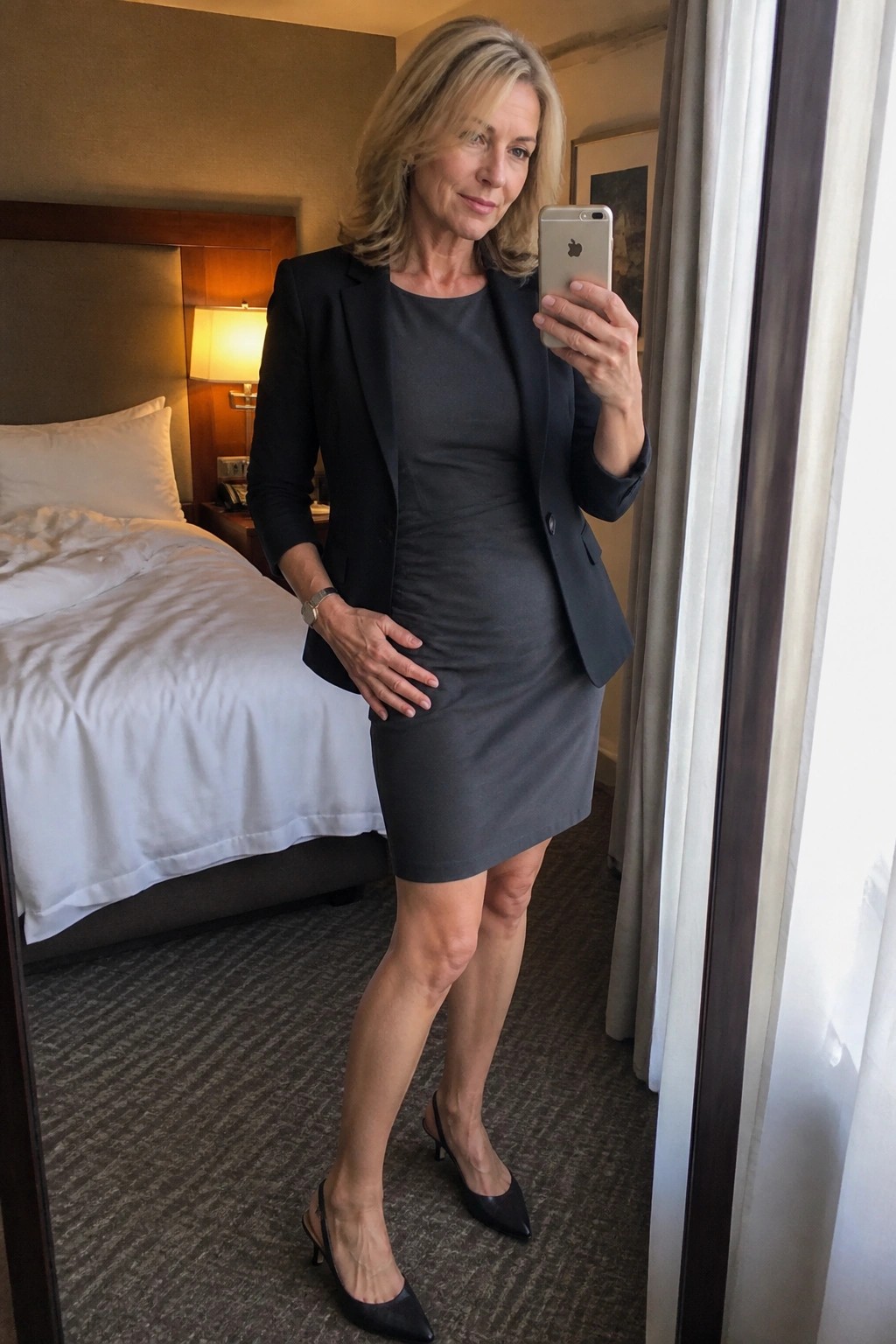 Woman in gray dress and black blazer taking mirror selfie in hotel bedroom