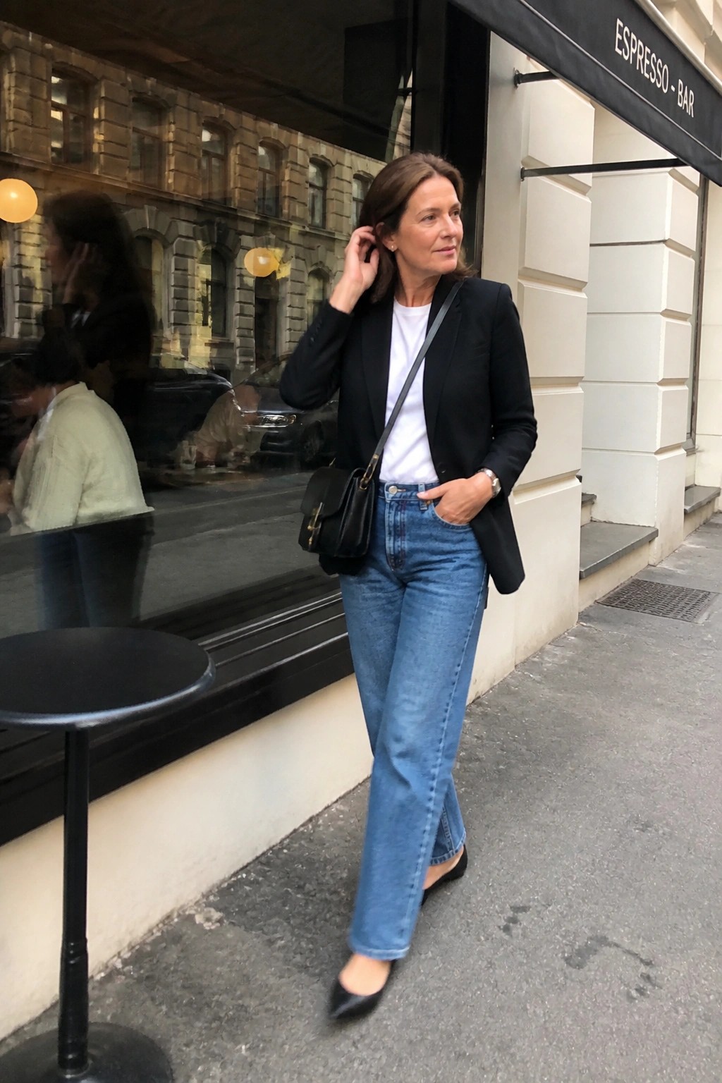 Woman in black blazer, white tee, and blue jeans outside espresso bar on sidewalk.