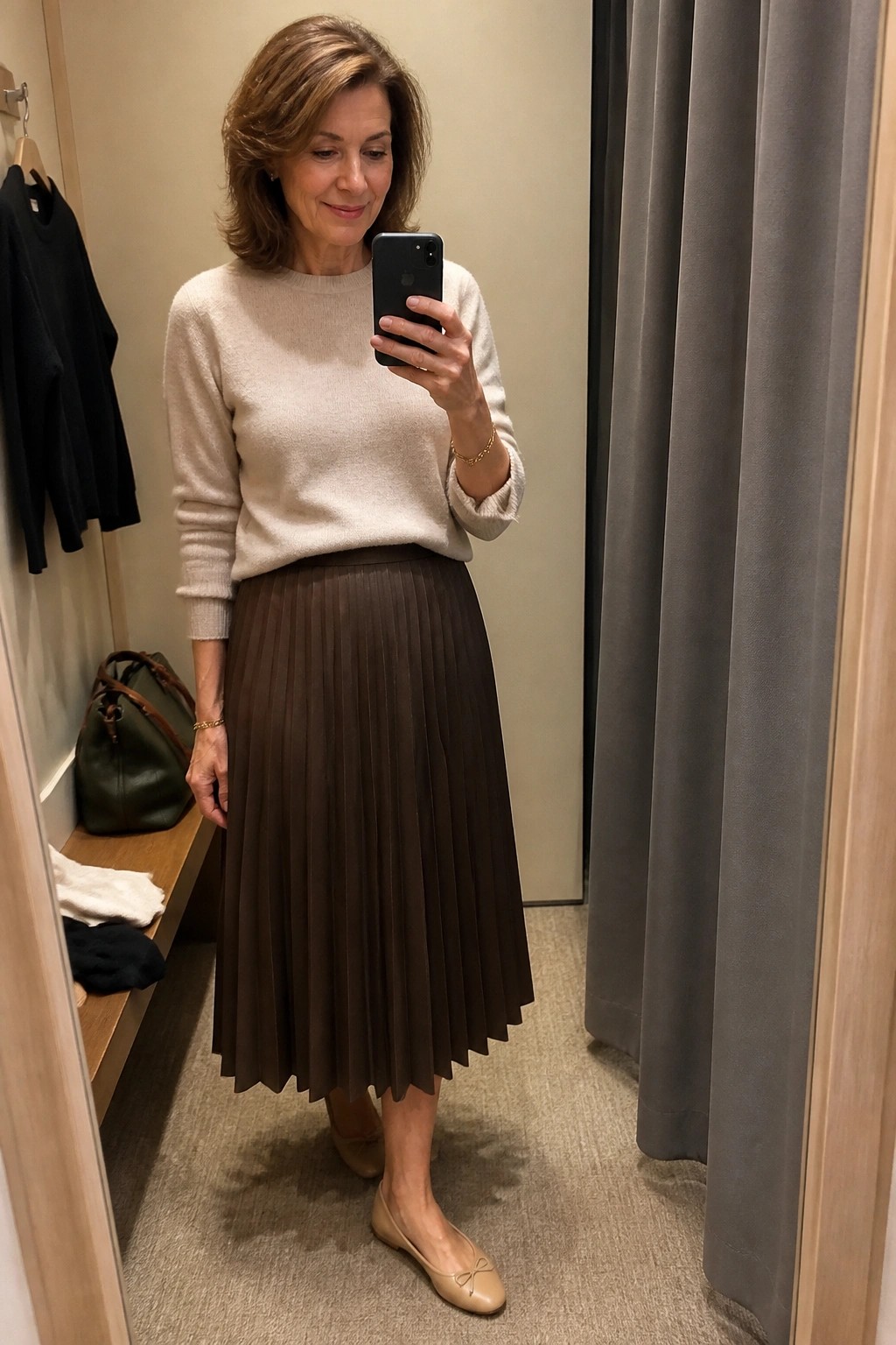 Woman in beige sweater and brown pleated skirt taking mirror selfie in fitting room