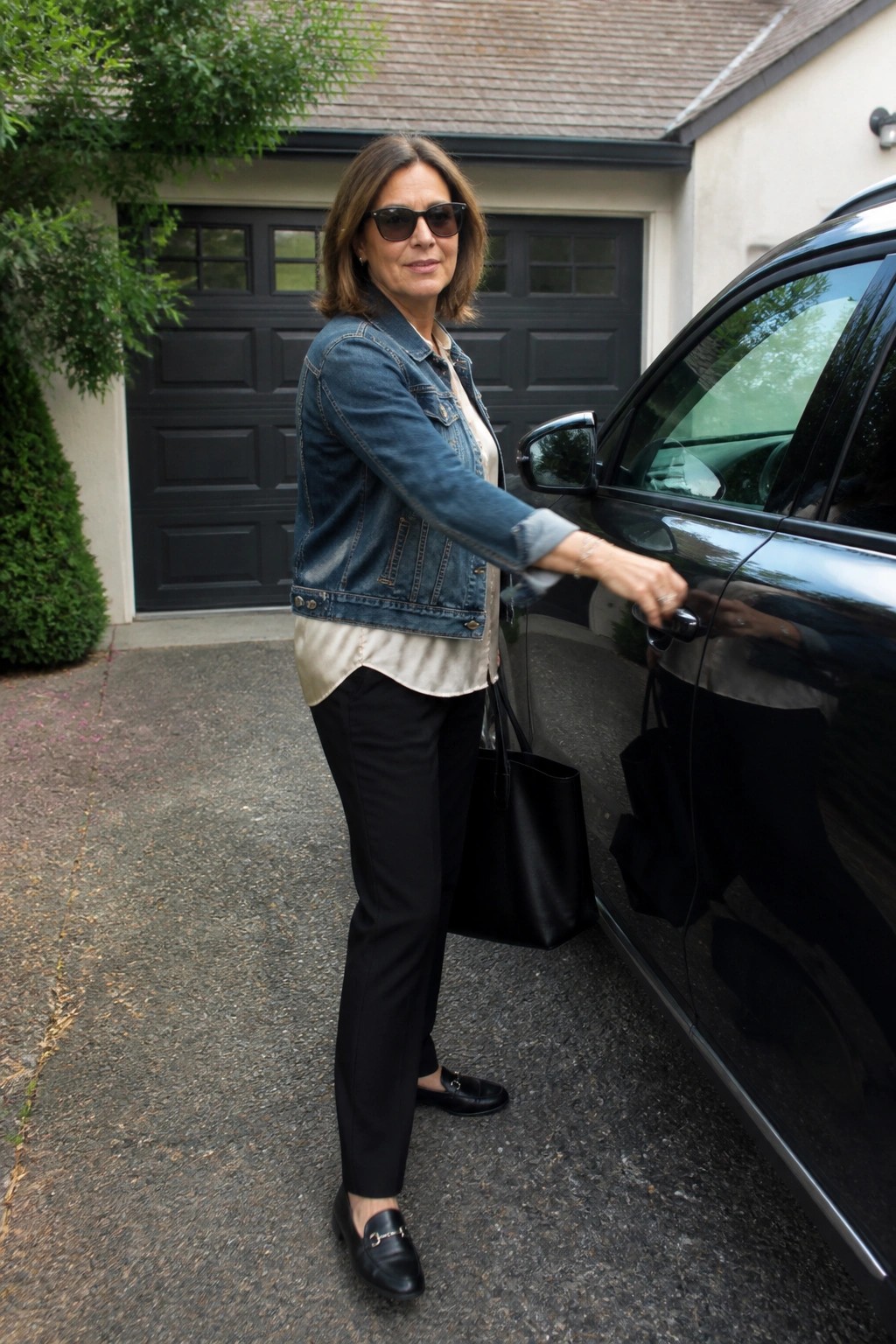 Woman in denim jacket and sunglasses holding black bag by shiny car door