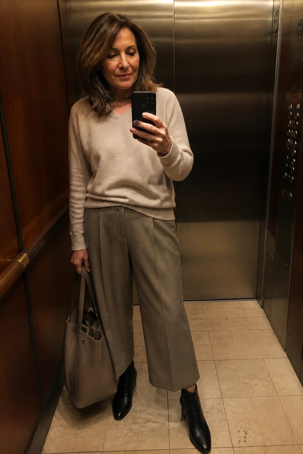 Woman in beige sweater and taupe pants taking selfie in elevator holding bag