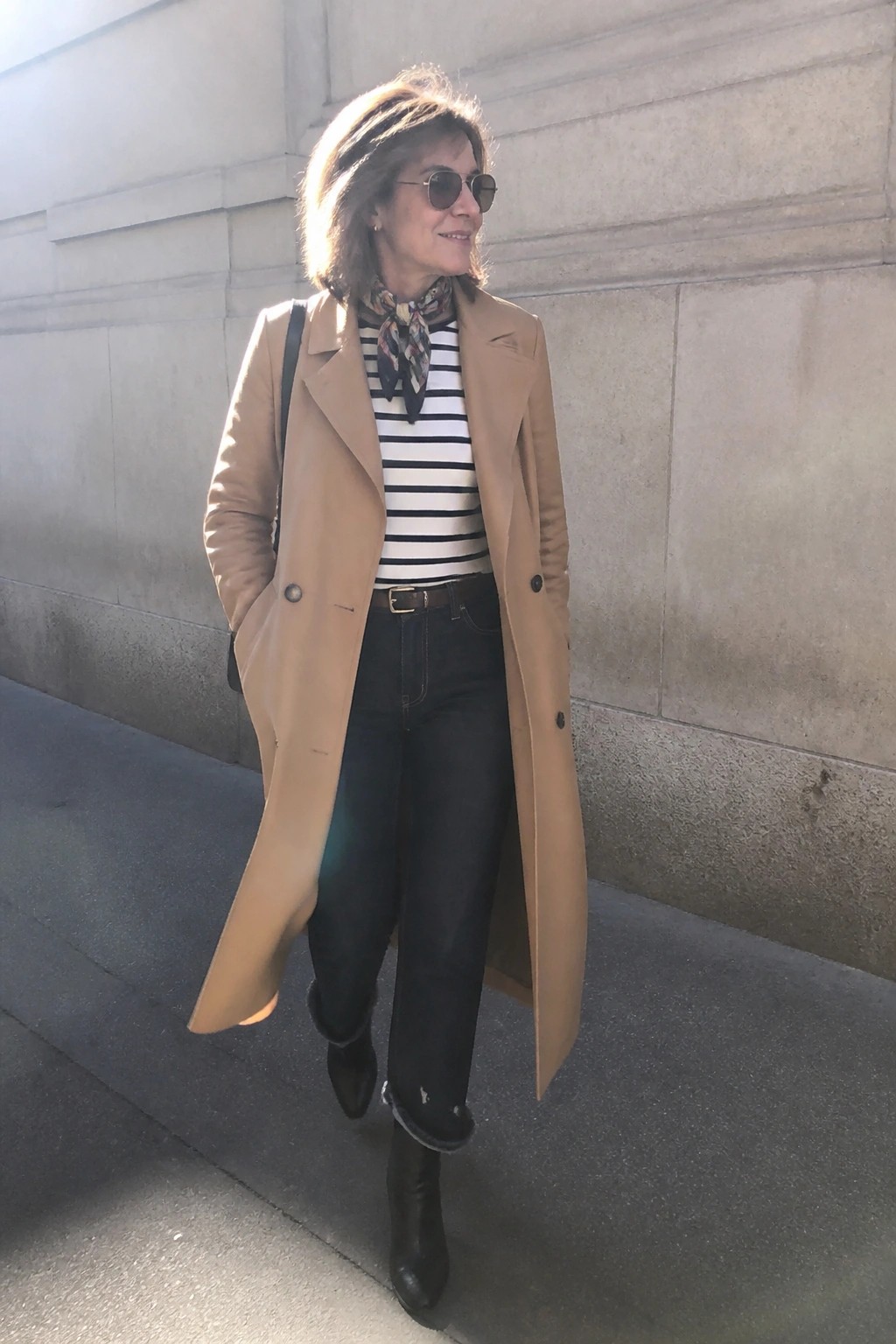 Woman in tan trench coat, striped top, scarf, and dark jeans by stone wall
