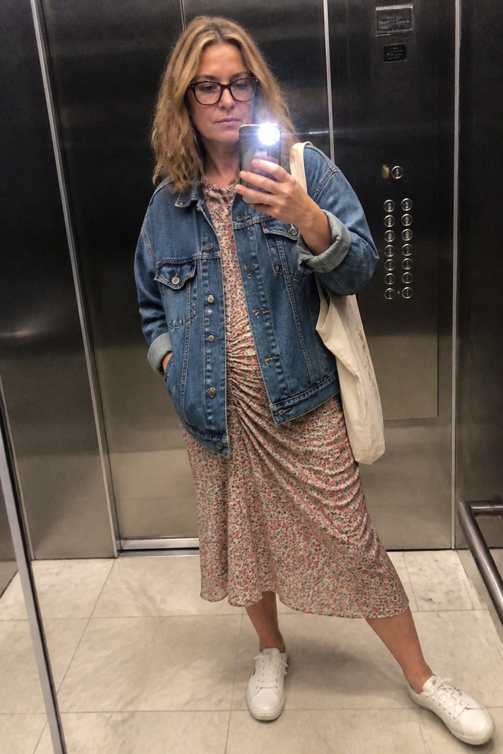Woman in denim jacket and floral dress taking mirror selfie in elevator